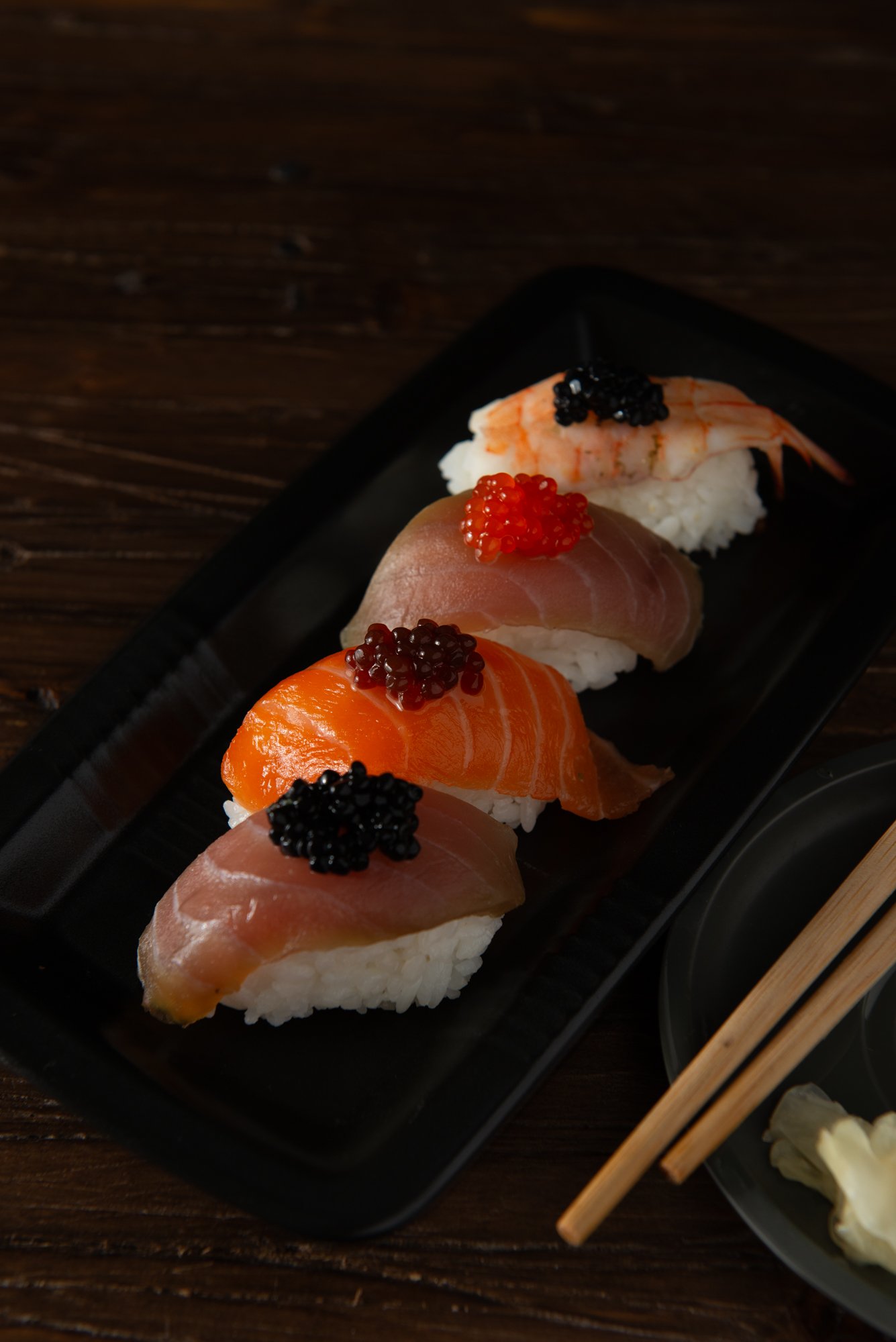 Conjunto de nigiri sushi con salmón, atún y camarón, adornado con huevas de pescado en un plato negro, acompañado de palillos japoneses.