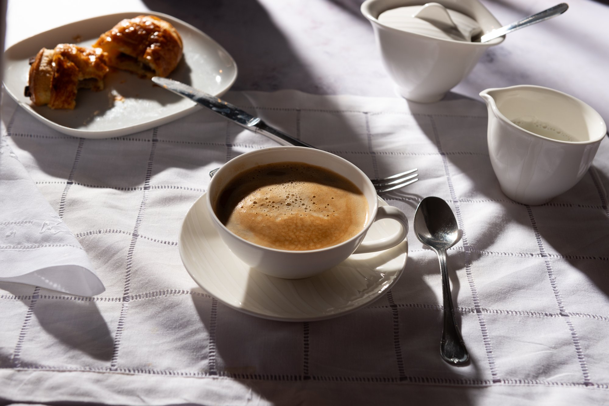 Taza de café sobre un mantel blanco con sombras, acompañada de un croissant mordido en un plato, una jarra de crema y una cucharita.