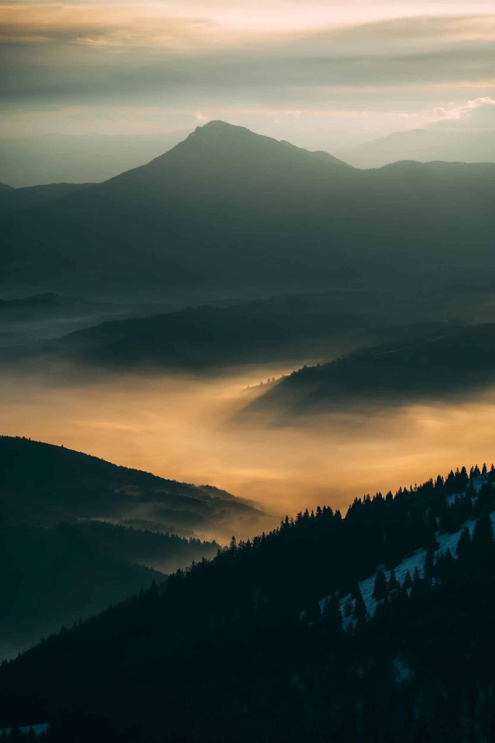 Layered mountain landscape during sunset or sunrise with fog in valleys and silhouetted trees on hills.