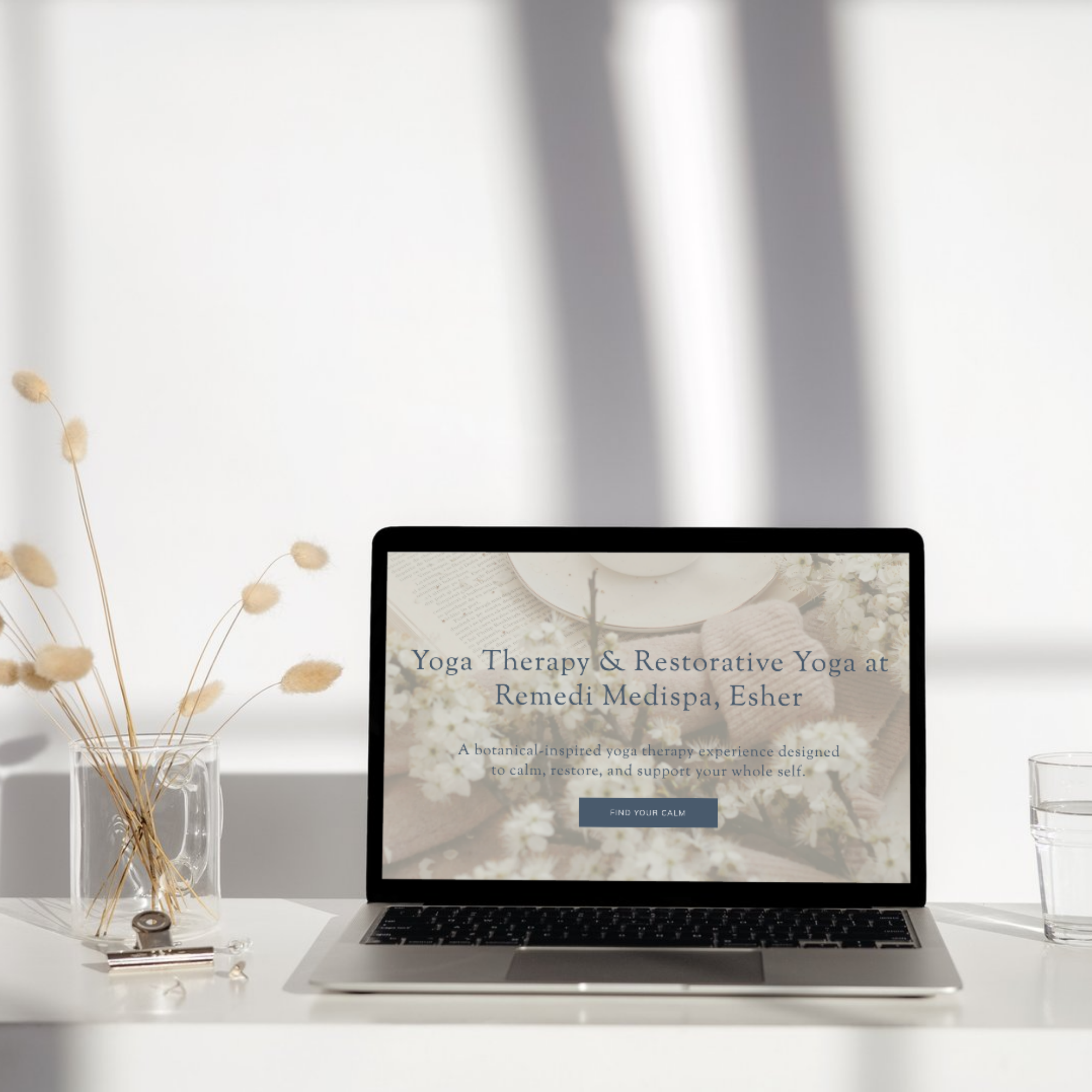 Laptop displaying a website about yoga therapy and restorative yoga at Remedi Medispa, Esher, on a white desk with a glass of water and a vase of dried flowers.