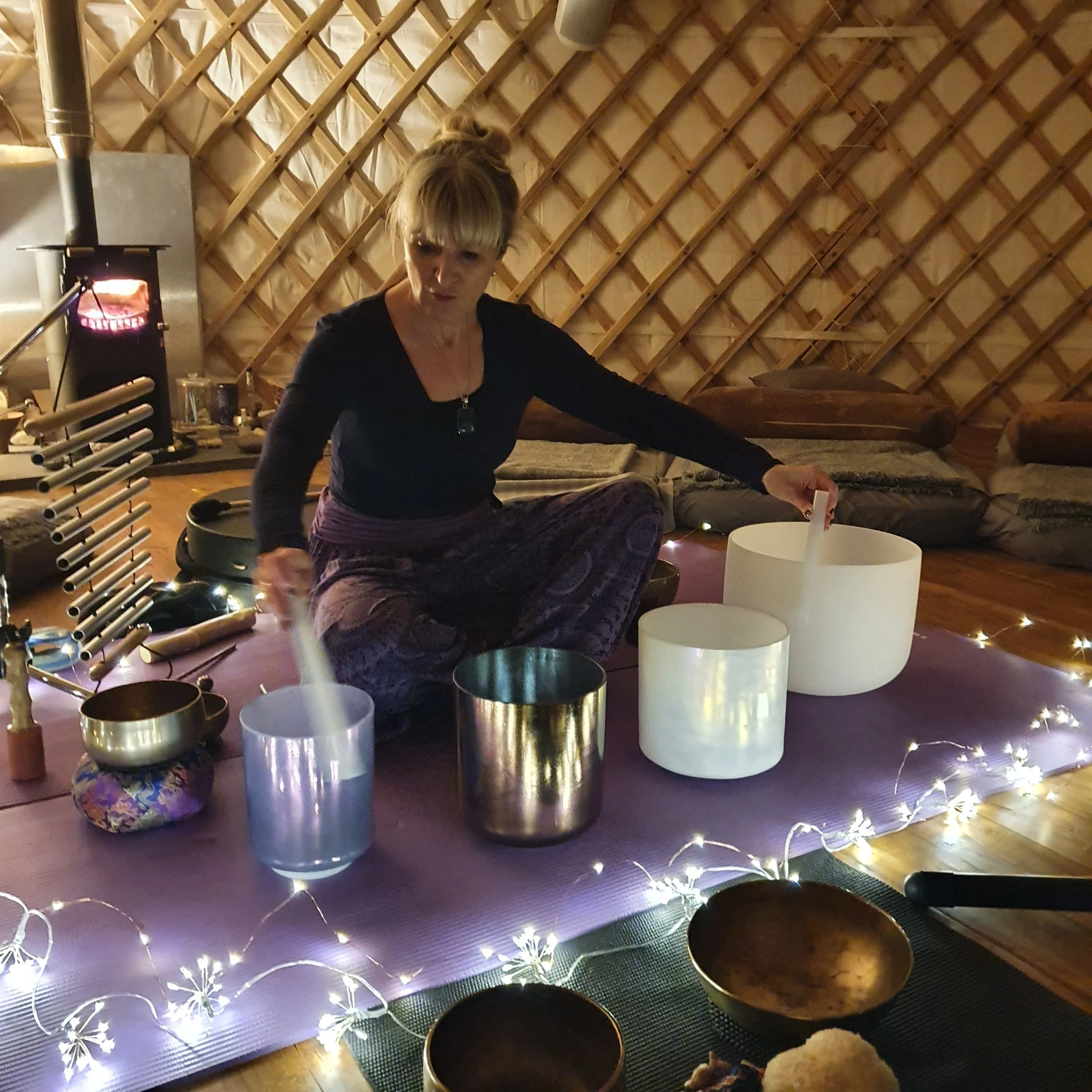 A woman sitting on a purple mat in a cozy, dimly lit yurt, playing crystal singing bowls surrounded by fairy lights and other musical instruments.