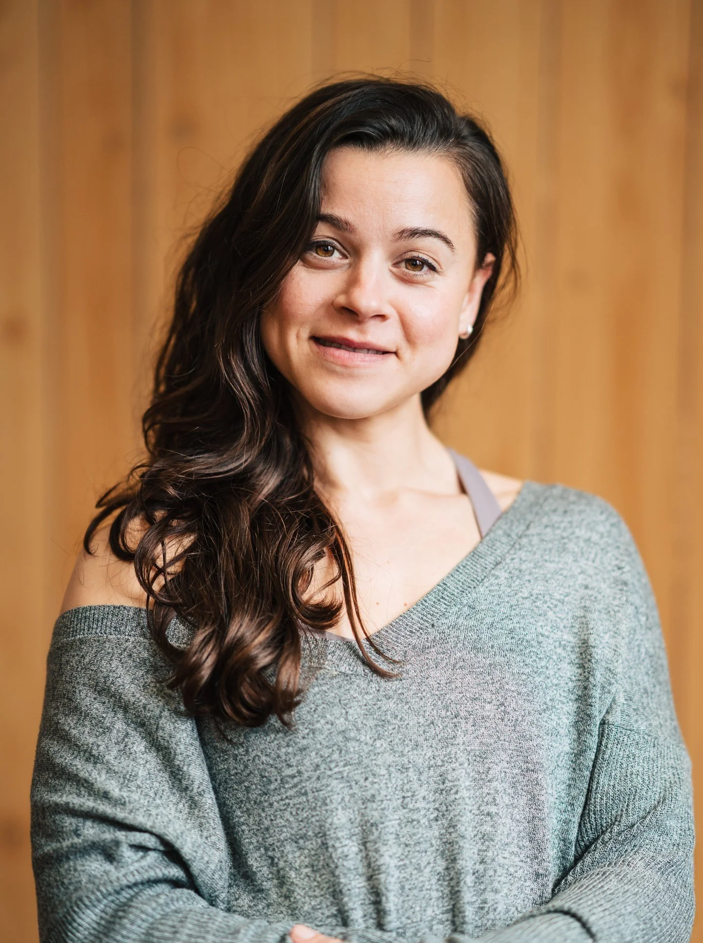 A woman with long, dark wavy hair wearing a gray sweater, smiling slightly, with a wooden background.