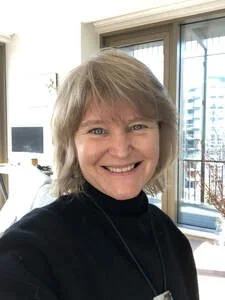 A woman with short, light brown hair and a black top smiling in an indoor setting, with a window showing a cityscape in the background.
