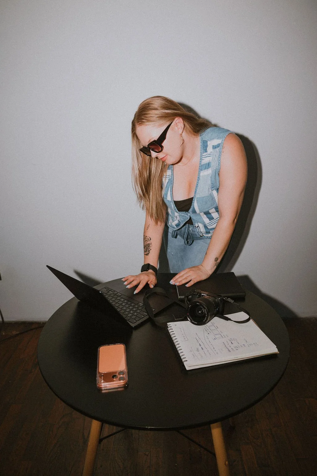 A person standing at a table and looking down at an open laptop beside an orange phone and notebook.