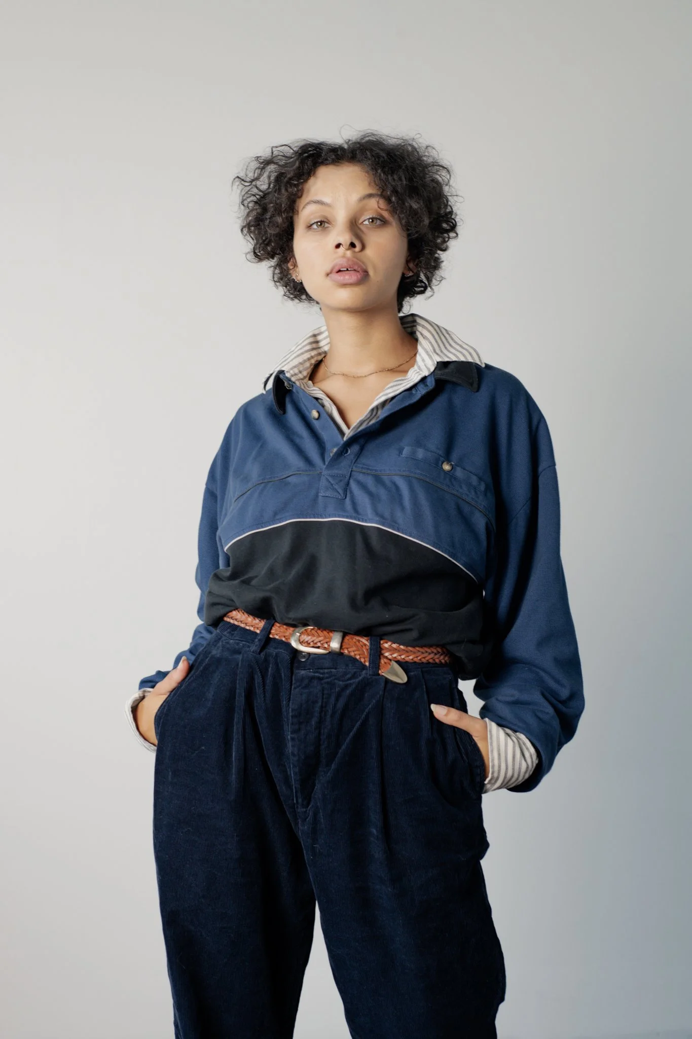 Studio fashion portrait of a model wearing dark blue pants, a two-toned windbreaker, and a brown woven belt against a clean white background.