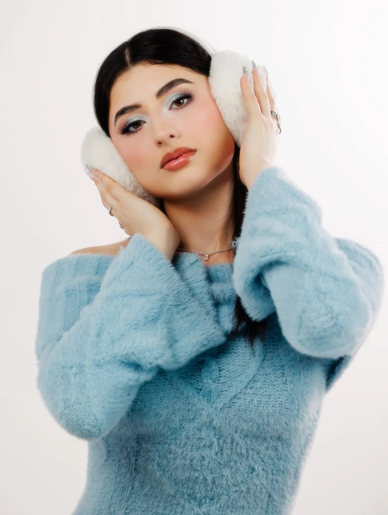 Studio fashion portrait of a model wearing a soft blue sweater and white earmuffs against a clean white background.