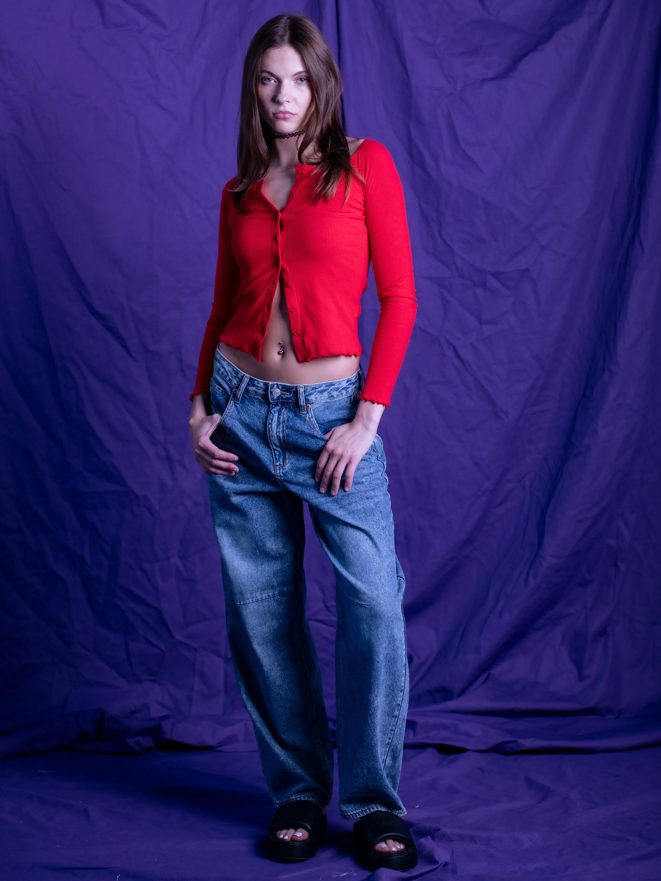 Full-body fashion portrait of a model wearing a red top and jeans against a purple backdrop.