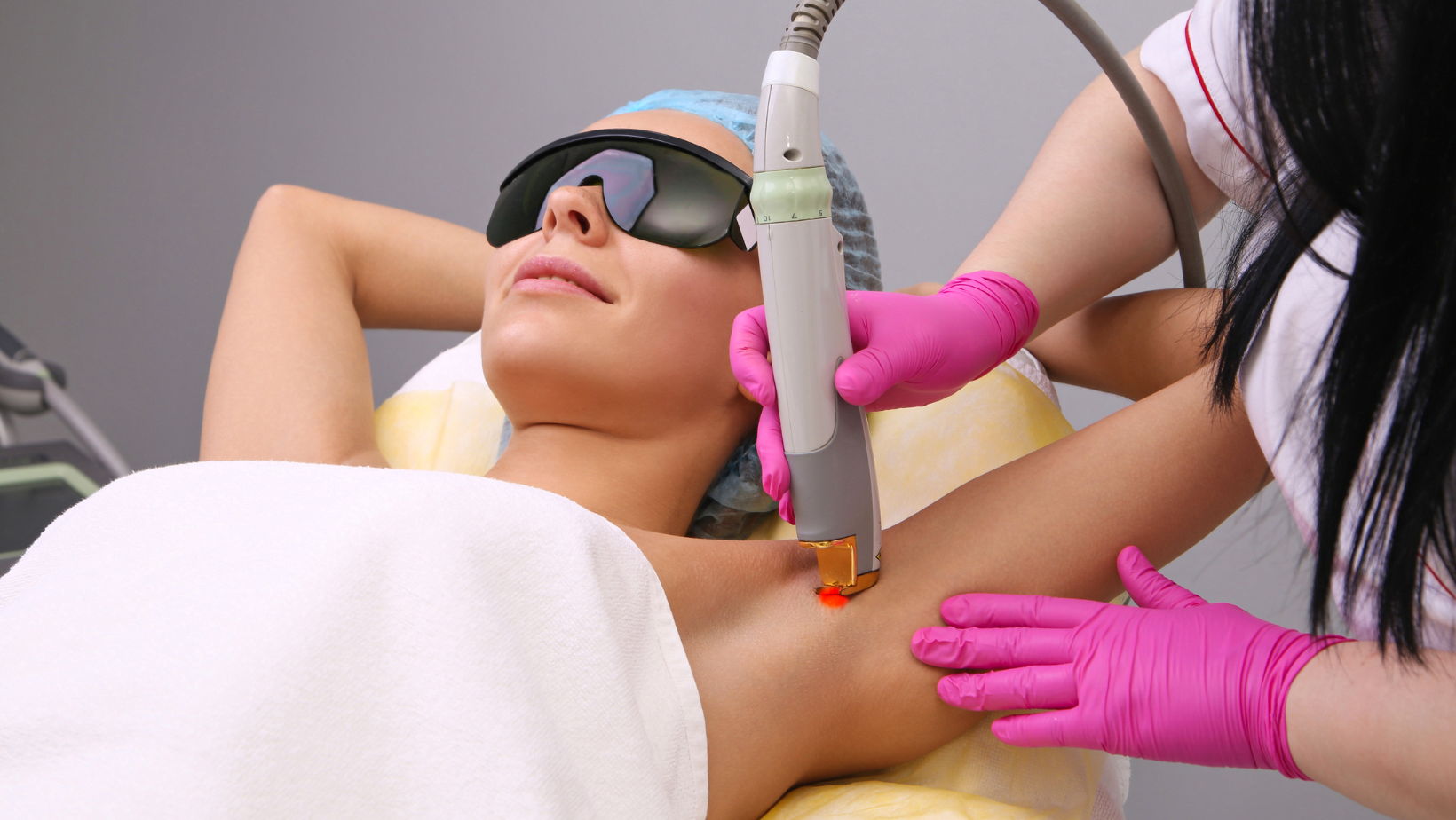 A woman lying on a medical bed with protective goggles, receiving laser hair removal treatment on her underarm by a technician wearing pink gloves.
