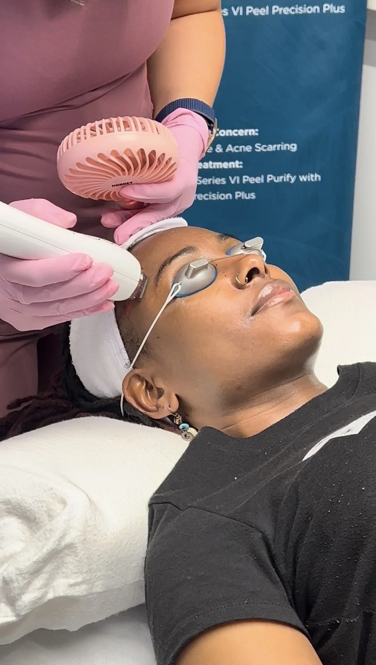 A woman with skin of color lying on a treatment bed receiving a Sciton Moxi skincare facial procedure, with a skincare technician holding a device. The woman is wearing protective goggles.