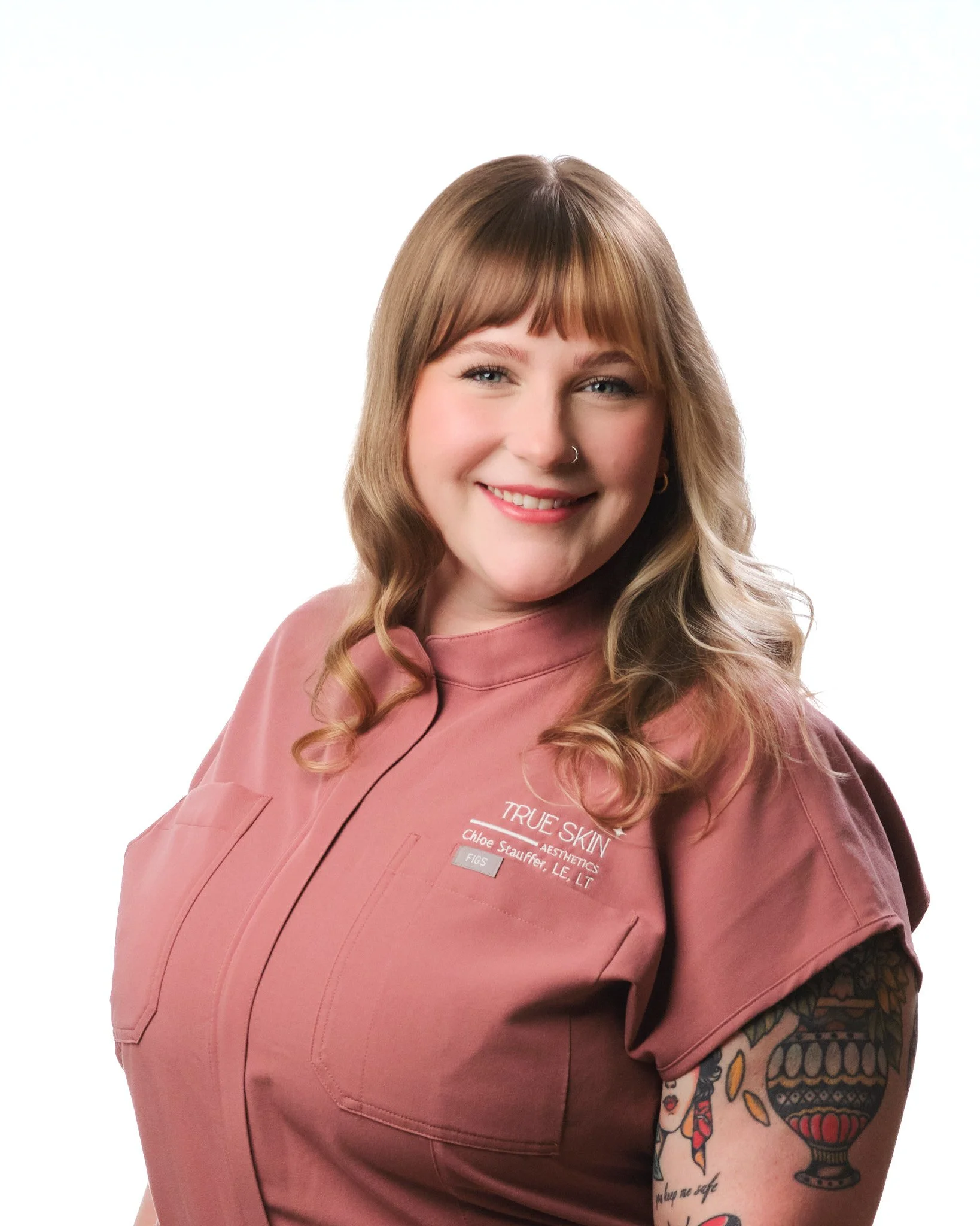 Close-up of a smiling True Skin Aesthetics team memeber with tattooed arm, wearing a pink work uniform with text on it, against a plain white background.