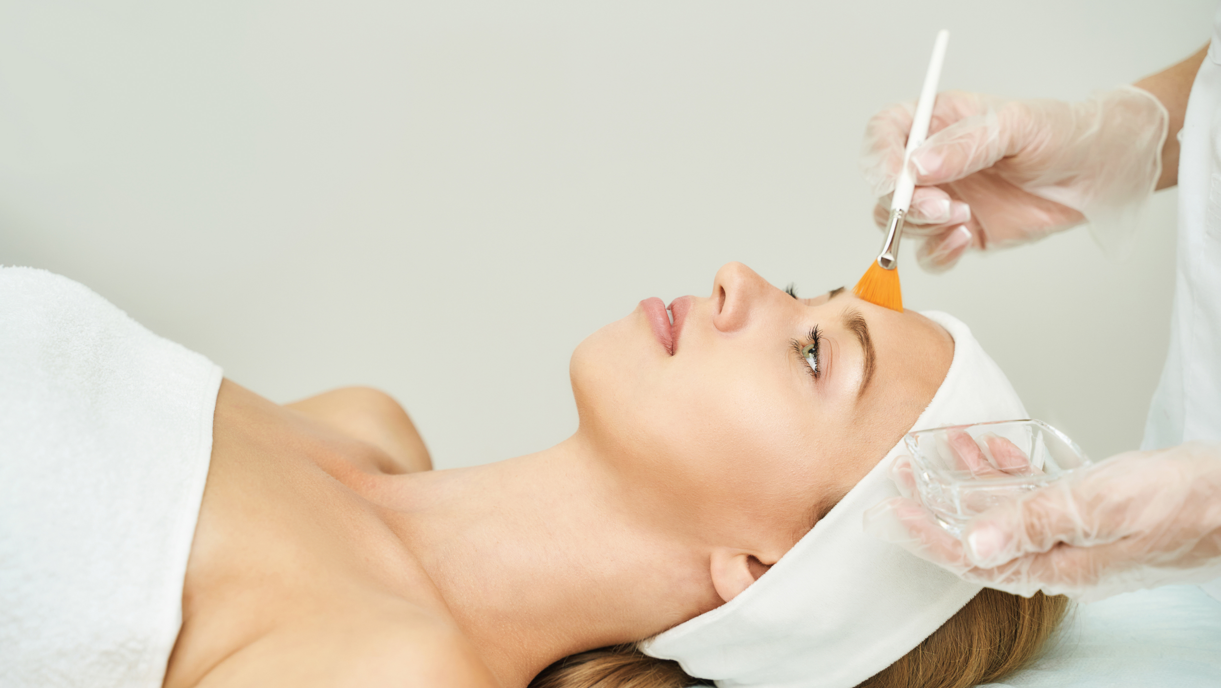 A woman lying down undergoing a facial treatment by a skincare professional wearing gloves, using a brush to apply a product near her face.