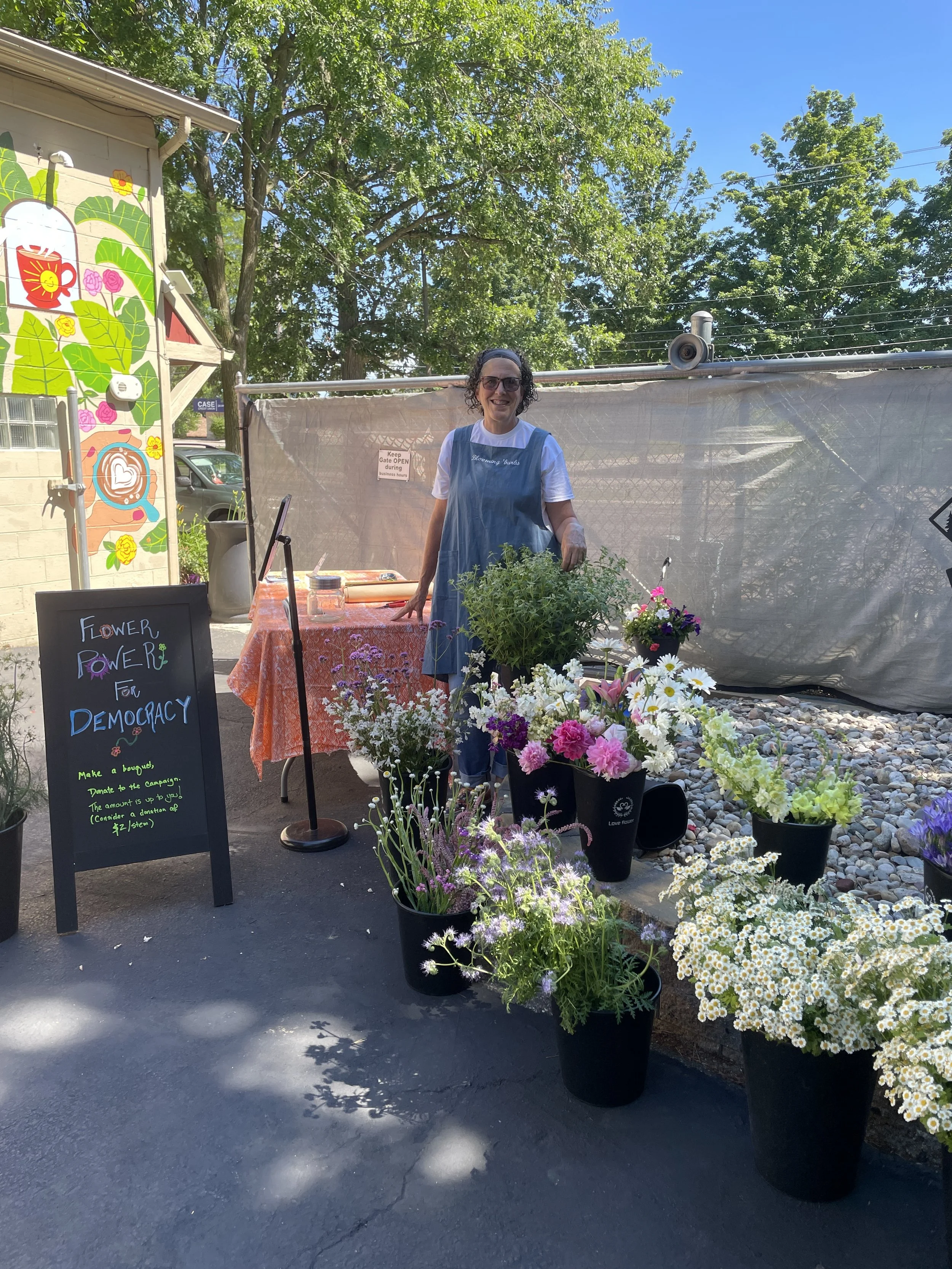 A white woman stands next to many buckets of flowers. The scene is outside, there is a blue sky and trees in the background, and also a wall painted with leaves and other colorful items.