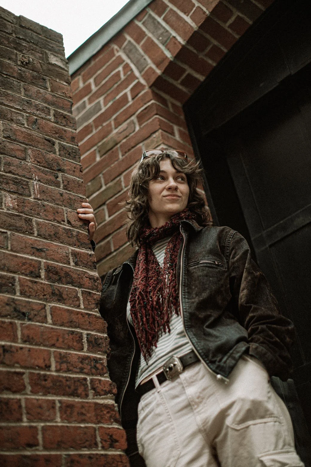 A young woman with curly brown hair stands next to a brick wall, wearing a dark jacket, a patterned scarf, and light-colored pants, looking confidently into the distance.