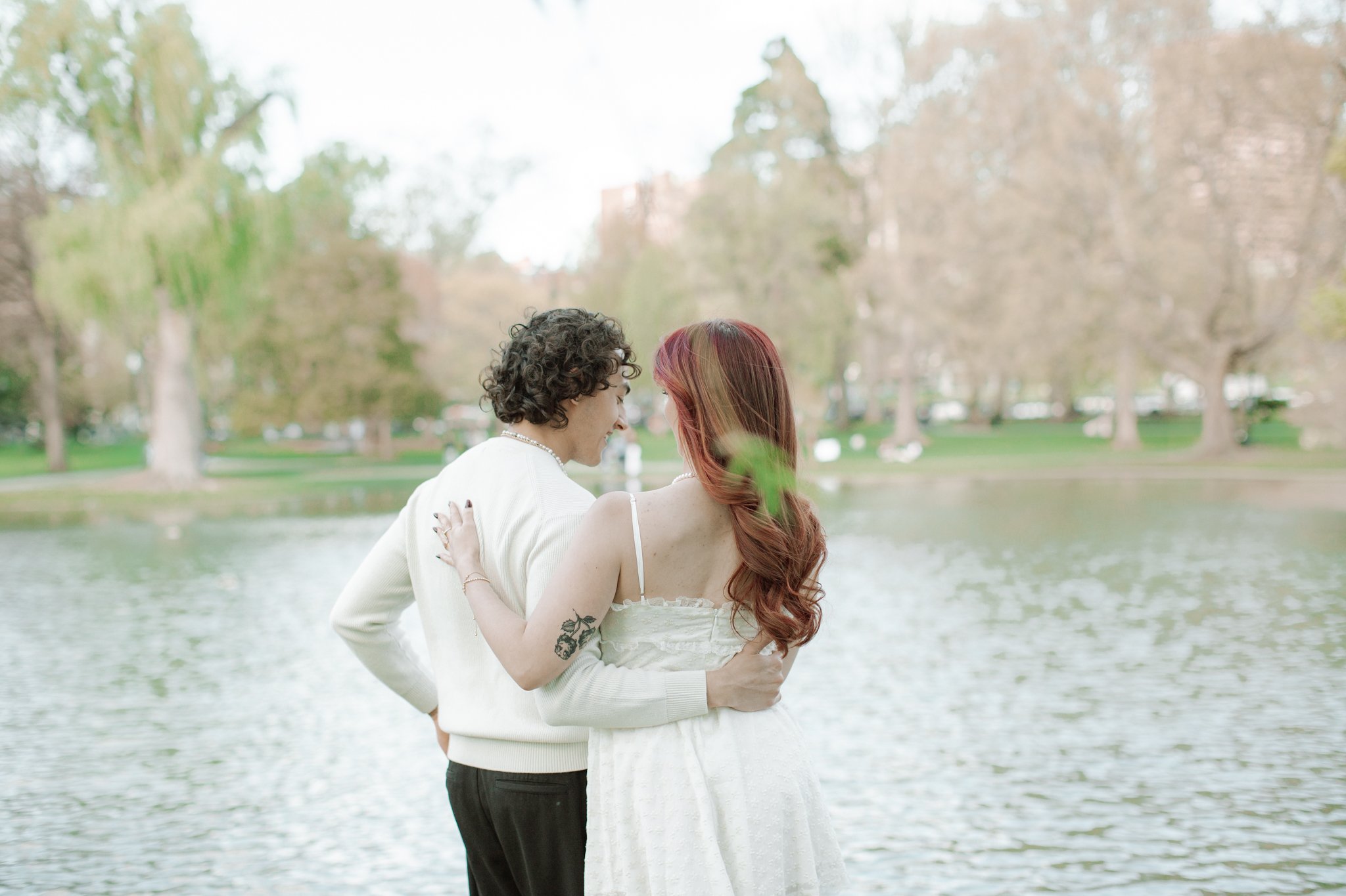 A couple dressed in wedding attire hugging by a lake in a park with trees in the background.