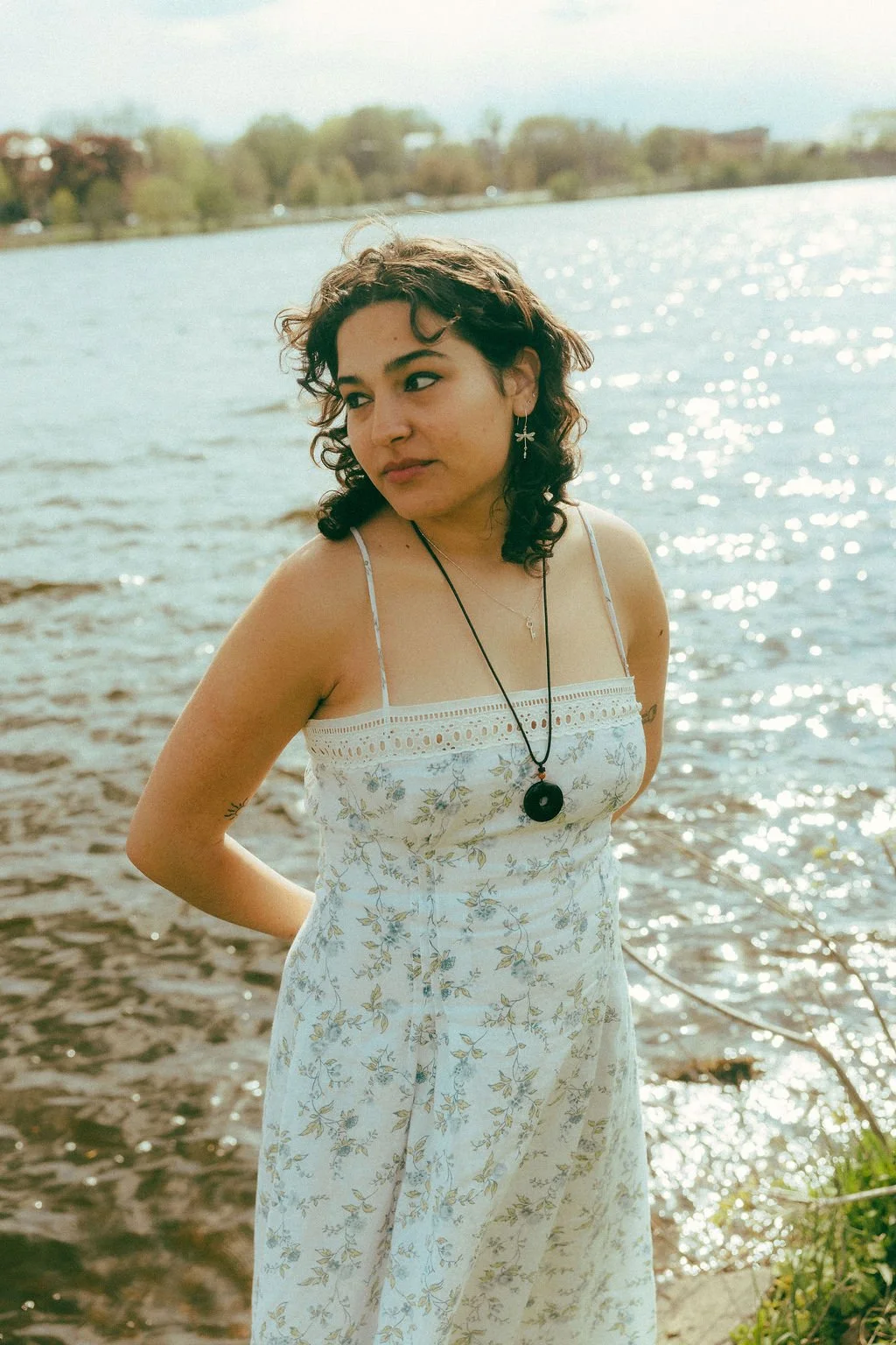 A woman standing by a body of water with trees and buildings in the background, wearing a white floral dress, with curly hair, earrings, and necklaces, looking to the side.