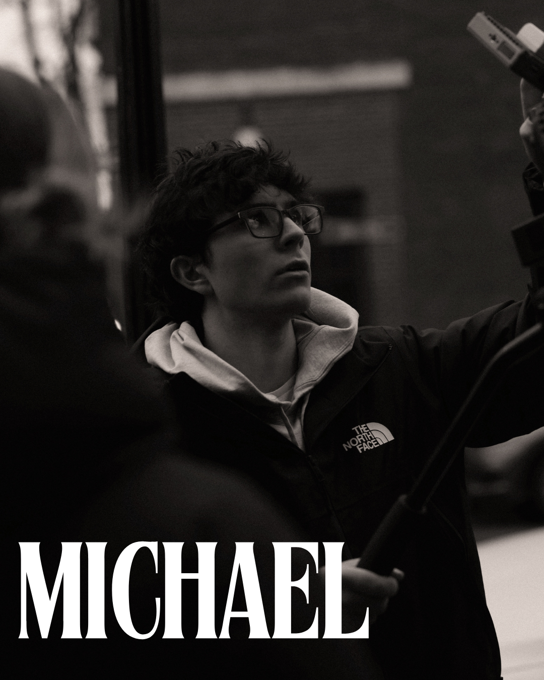 A young man with curly hair and glasses, wearing a hoodie and a North Face jacket, is looking up and appears to be working with equipment or a camera in a dimly lit indoor setting.