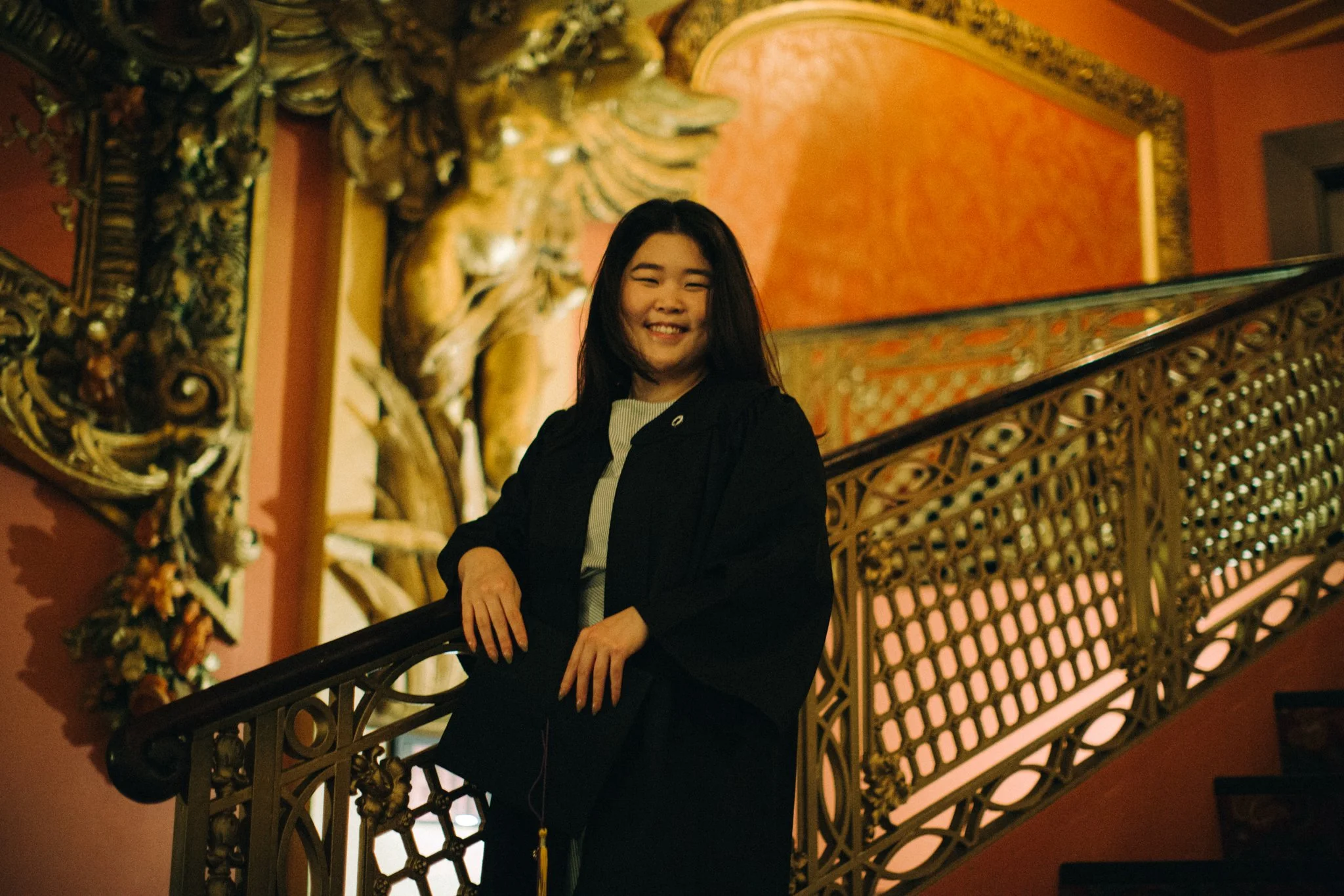 A young woman with black hair smiling, standing on an ornate staircase with intricate gold and wood details, against a pink and gold decorated wall.