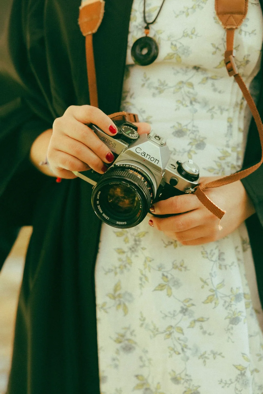 Person holding a vintage Canon film camera with a brown strap, wearing a floral dress and a black jacket, with red nail polish.