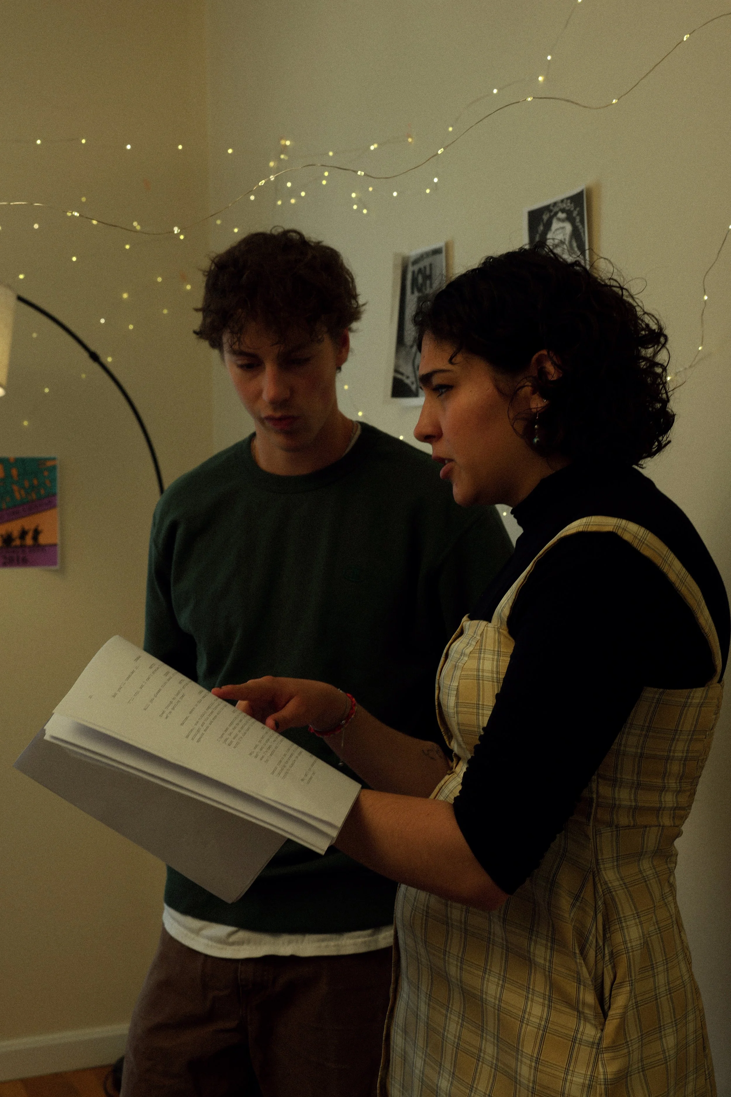Two young adults, a man and a woman, standing close together in a dimly lit room with string lights hanging from the ceiling. The woman holds an open book while both look at it attentively.