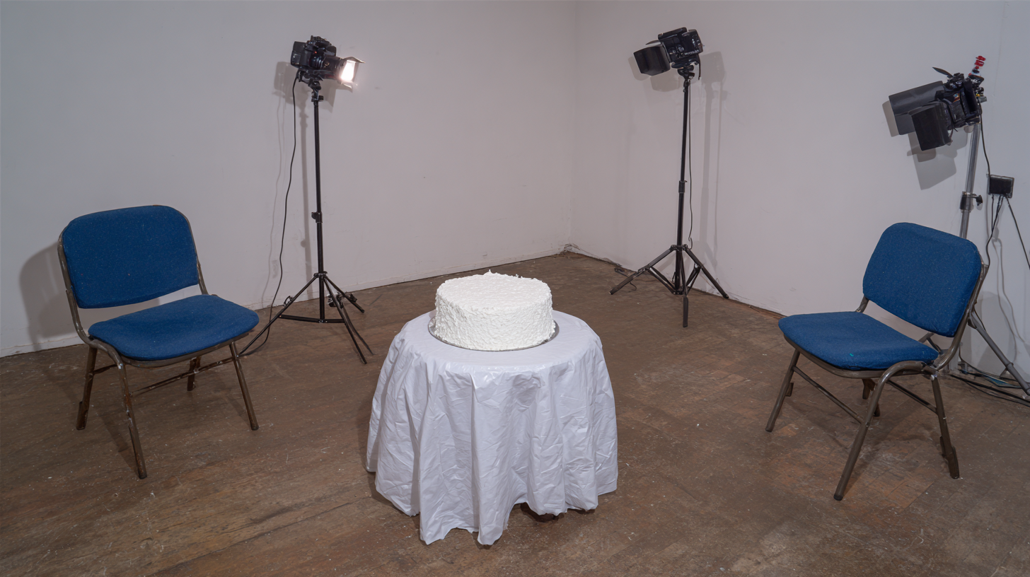 En hvid kage på et bord med en hvid dug, omgivet af tre blå stole og fire professionelle lampe- og kameraudstyr til fotografi eller videooptagelse.