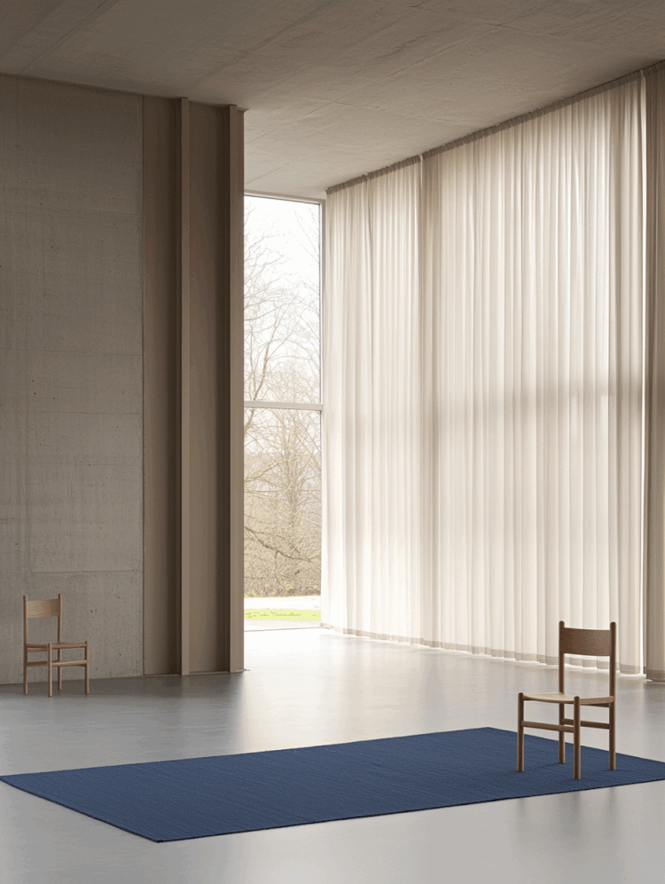 Minimalistisk værelse med store vinduer, to træstole og en blå måtte på gulvet. Gardiner hænger ved vinduerne.