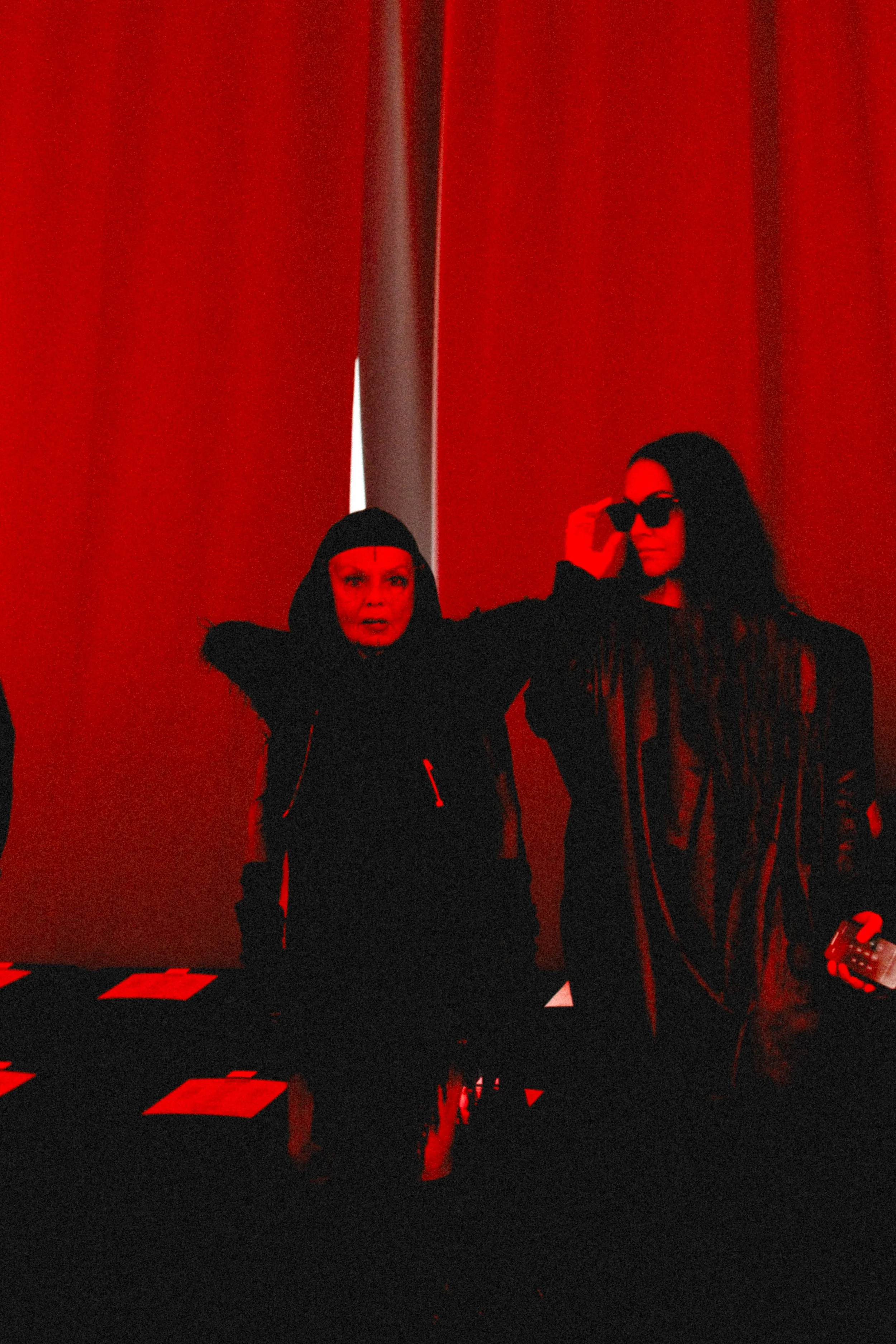 Michele Lamy and Christelle Caffarelli, studio rö's founder with red lighting and curtains in the background, one woman in black with sunglasses adjusting her glasses, the other woman in black with a hood, with papers on the table.