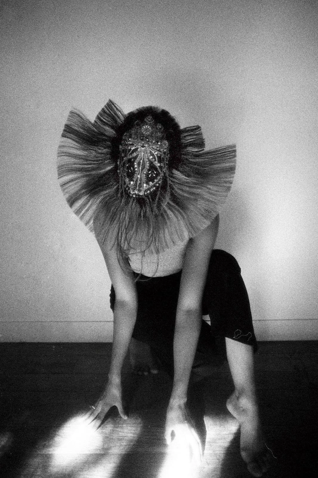A person crouching on the floor with each hand on the ground, one knee bent, wearing a lion mask with a decorative face and a large, ruffled collar, in a black and white photo.
