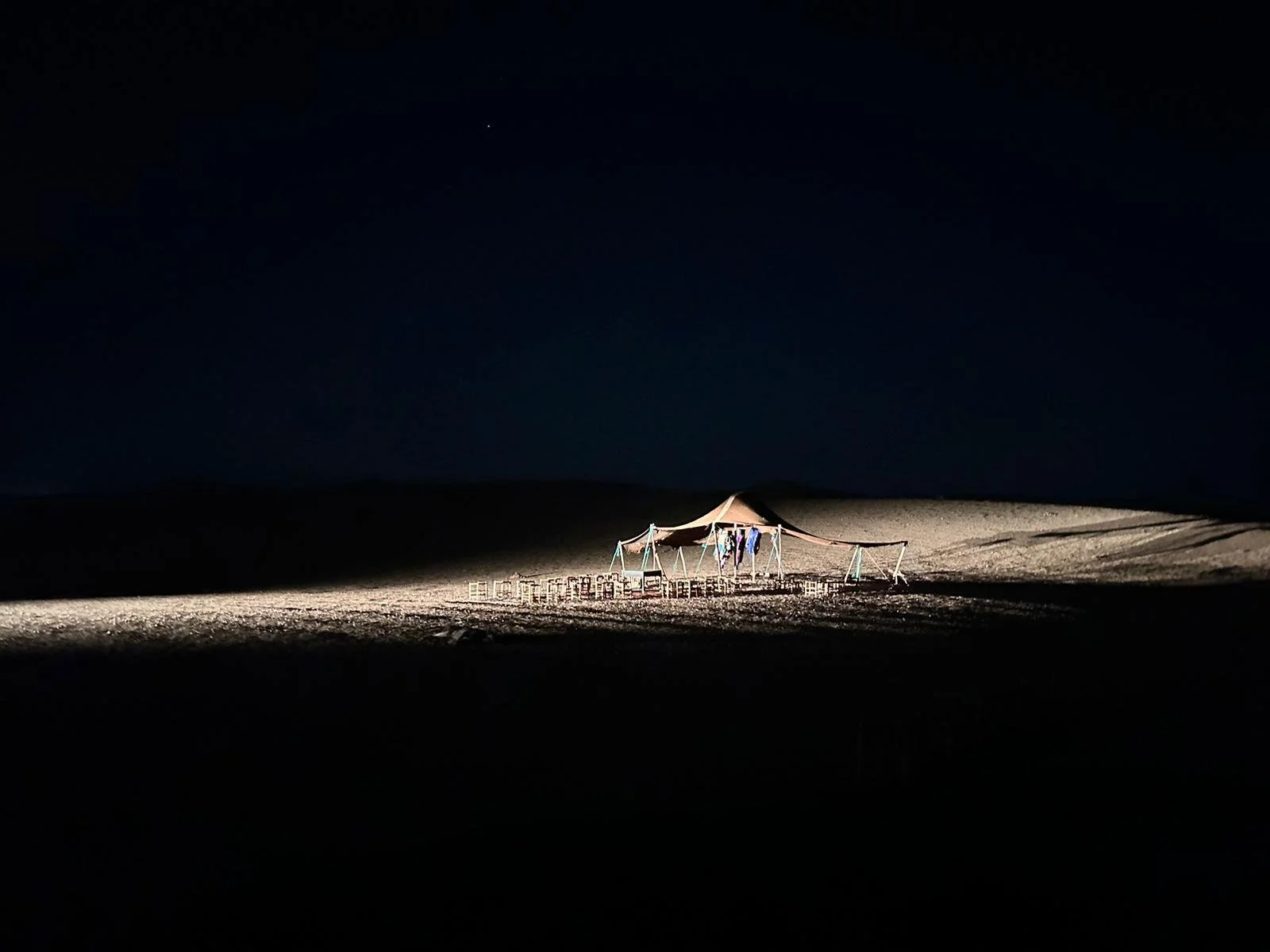 A tent set up in a desert at night with some clothes hanging inside, illuminated by a light, with a dark sky and faint stars visible.