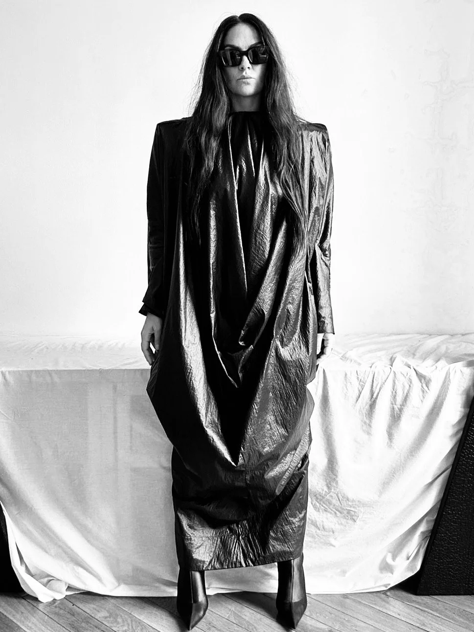Christelle Caffarelli, studio rö's founder, shiny, oversized dress with long sleeves, standing indoors against a white wall and a bed with a white sheet. She has long wavy hair, wears large black sunglasses, and black heeled boots.