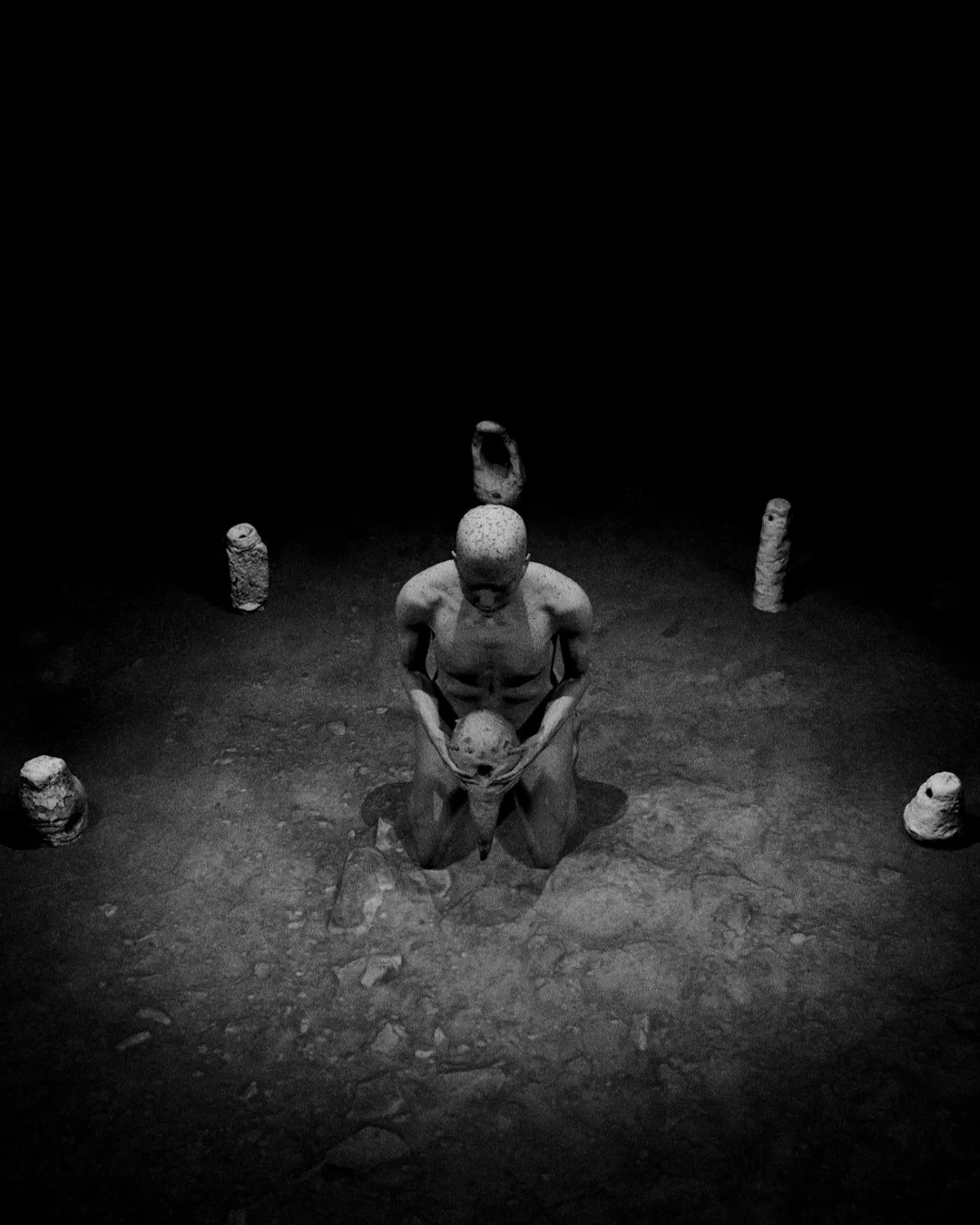 Black and white photo of a child statue kneeling and holding a skull, surrounded by six stone-like objects in a dimly lit space.