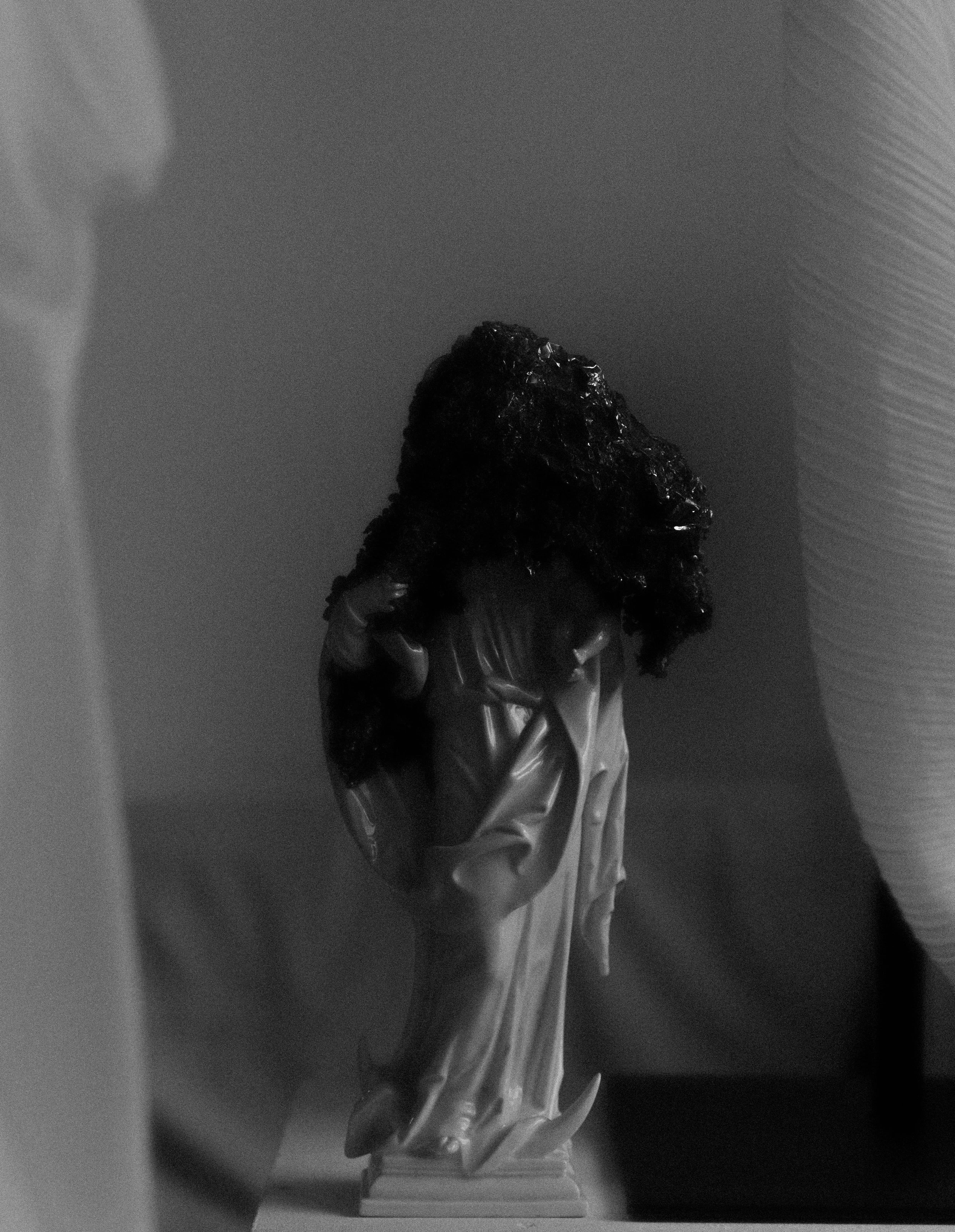 DAVID CLARKE AT STUDIO RÖ : Black and white photo of a statue with long, textured hair, wearing a long garment, standing on a pedestal, against a plain background.