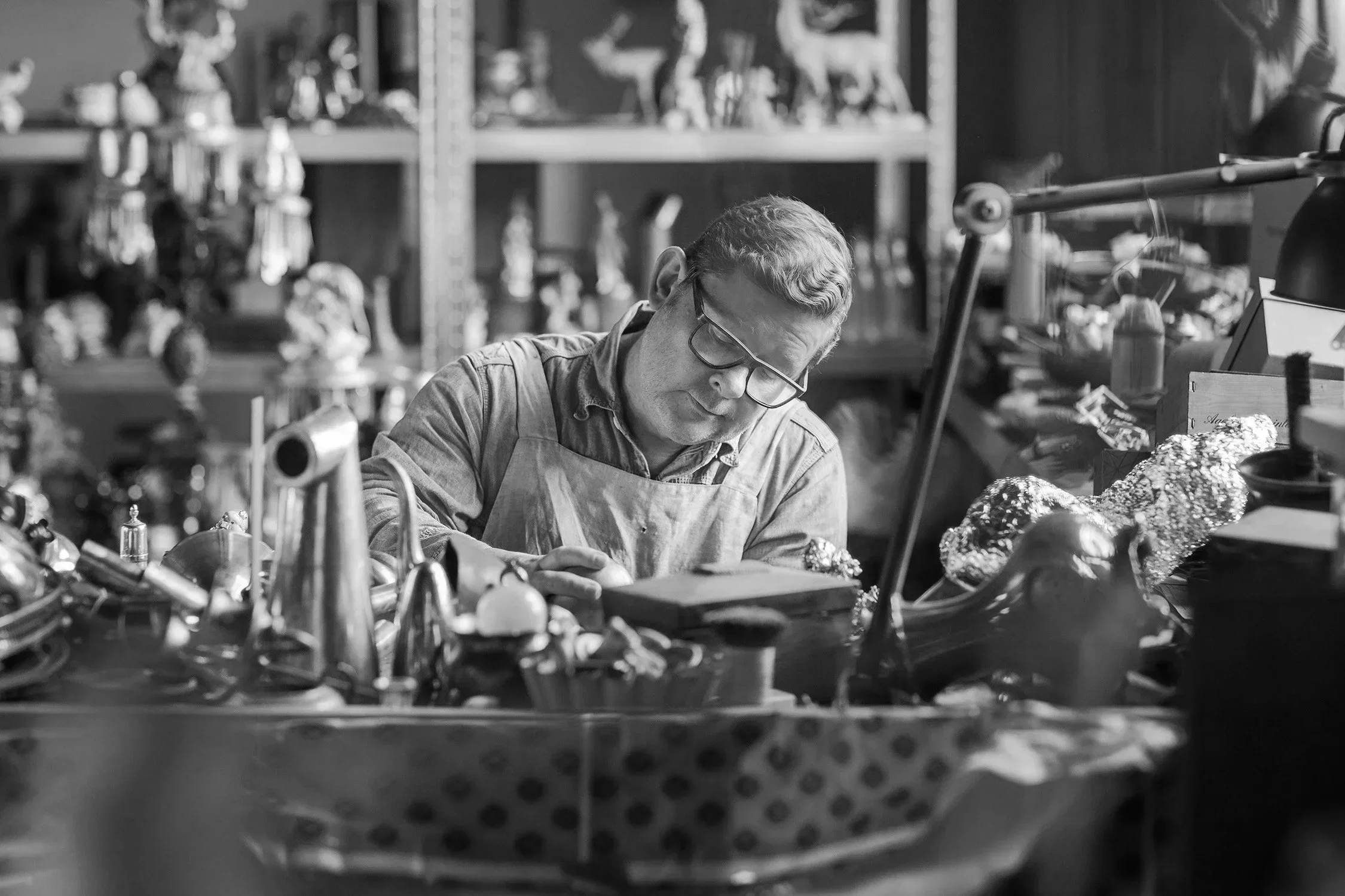 A man working at a cluttered workspace filled with various objects and tools, wearing glasses and an apron, with shelves of figurines and collectibles in the background.