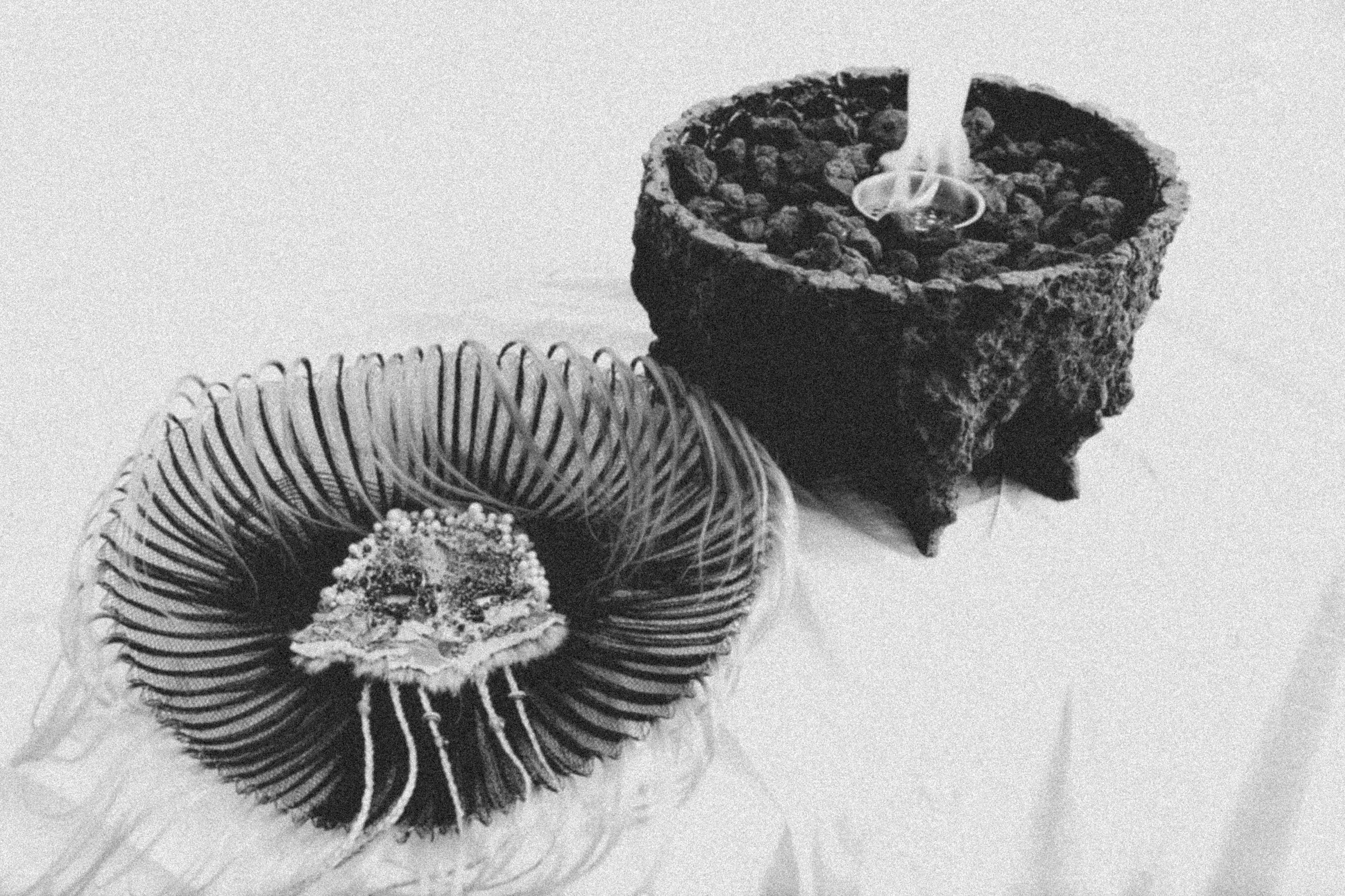 A black and white photograph of two candles in textured holders. The candle on the left is in a round, wire-like holder, and the candle on the right is in a rough, stone-like holder with melting wax on top.