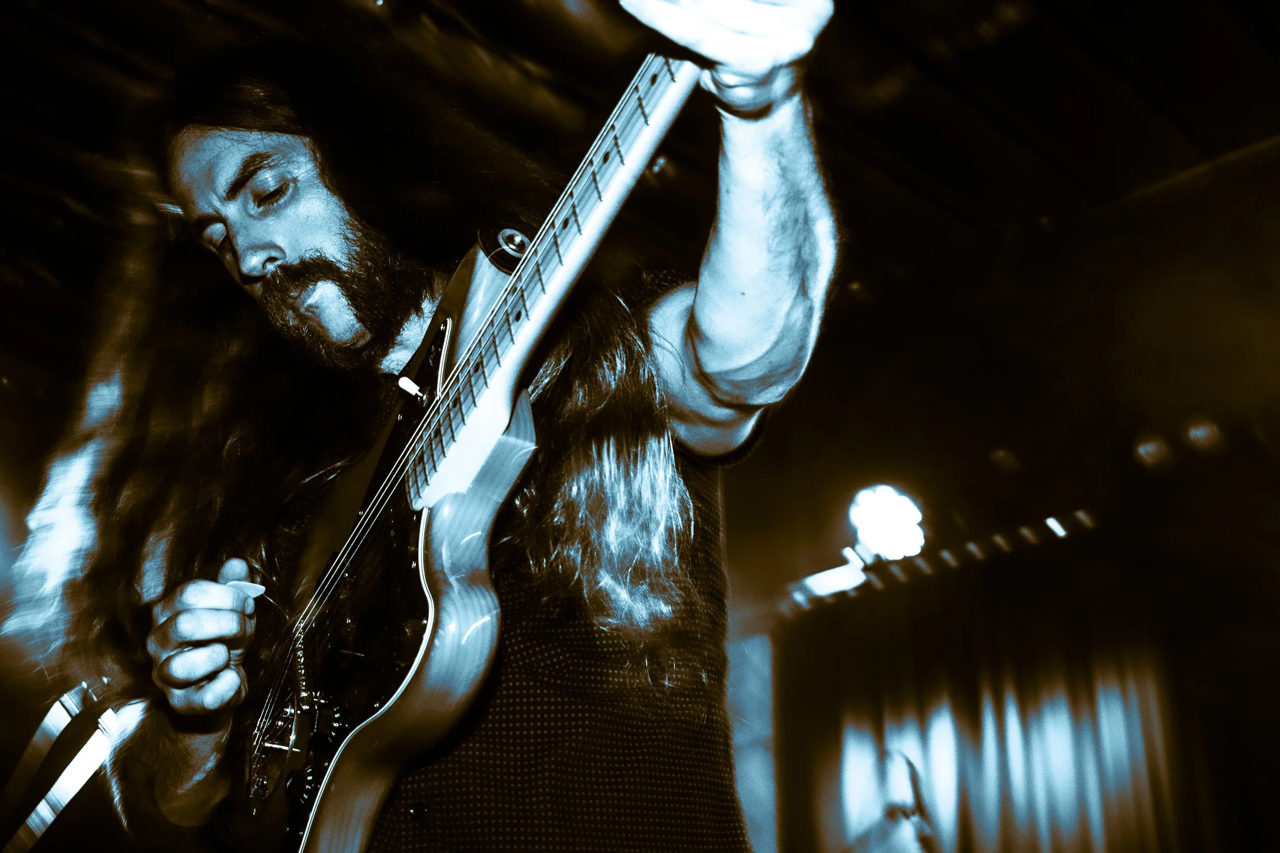 Rock band Liquid Pennies frontman/guitarist Chas BInns playing an electric guitar on stage, captured in blue-tinted lighting.
