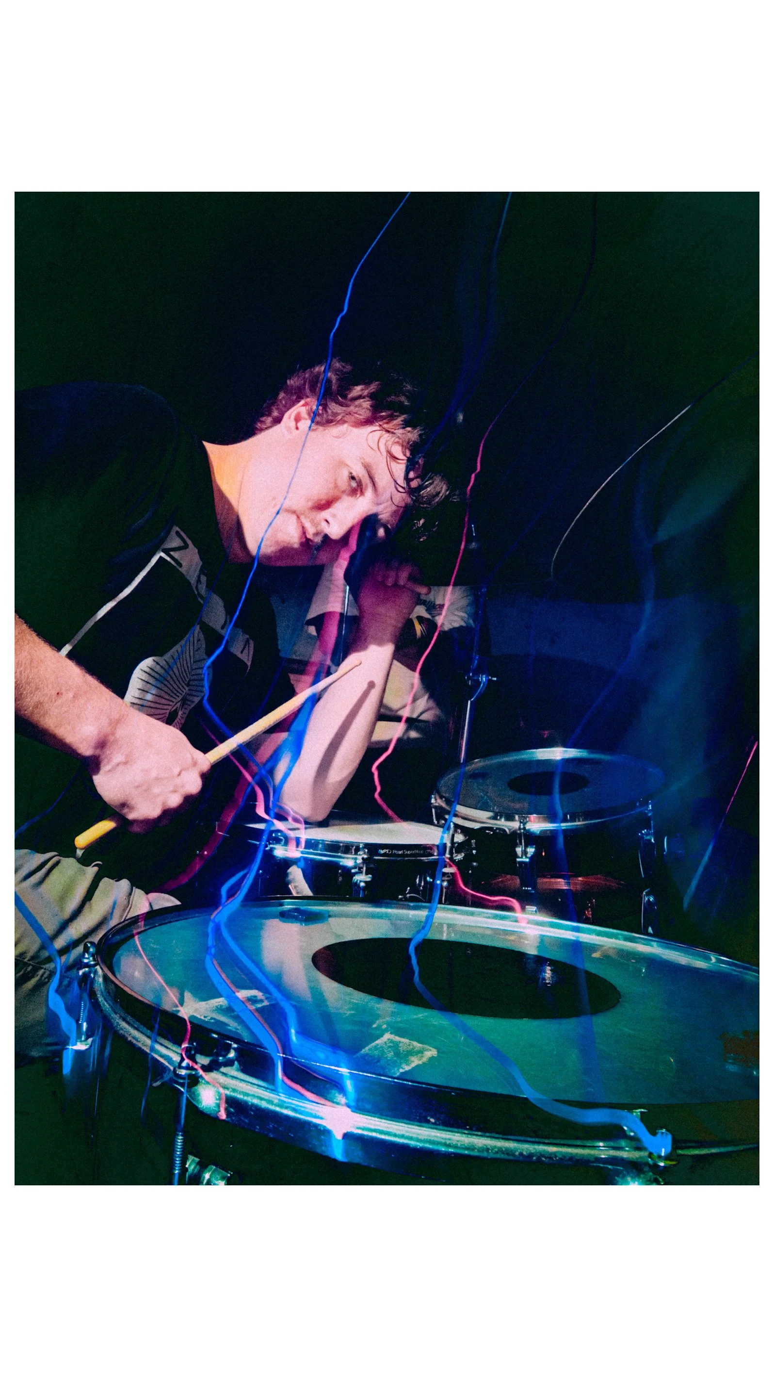 A person with curly hair and a black t-shirt playing drums, with colorful light streaks around him, in a dark setting.