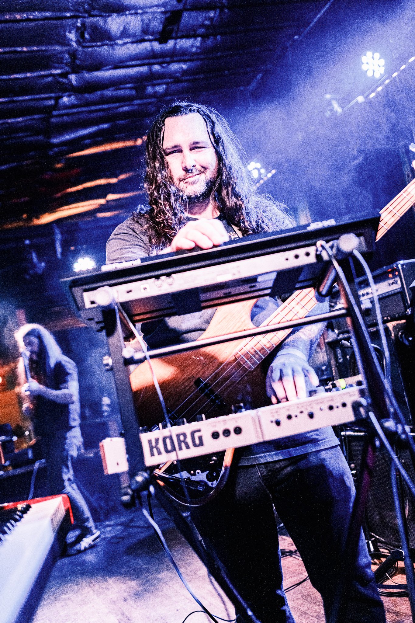 Tysonious Mink of the band Liquid Pennies  with long hair and a beard smiling at a concert stage, playing a bass guitar and standing behind a keyboard with a Korg logo, with stage lights and a dark indoor setting.