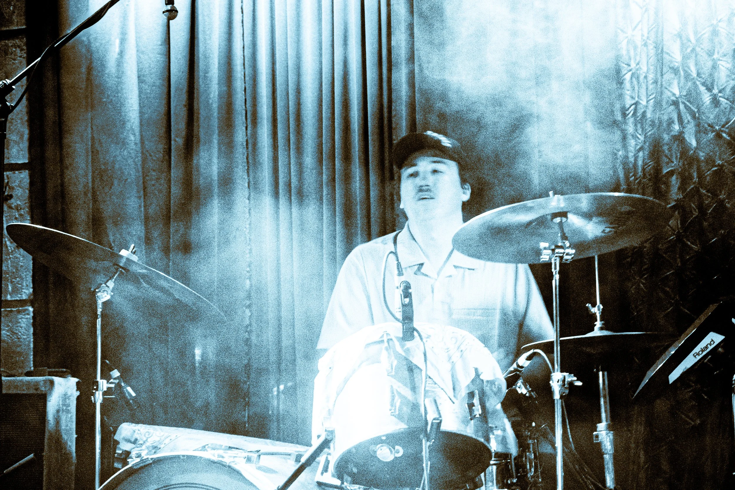 Indie rock band Liquid Pennies drummer PIerson Whicker  sitting behind a drum set, wearing a shirt and a cap, on a stage with curtains and stage lights.
