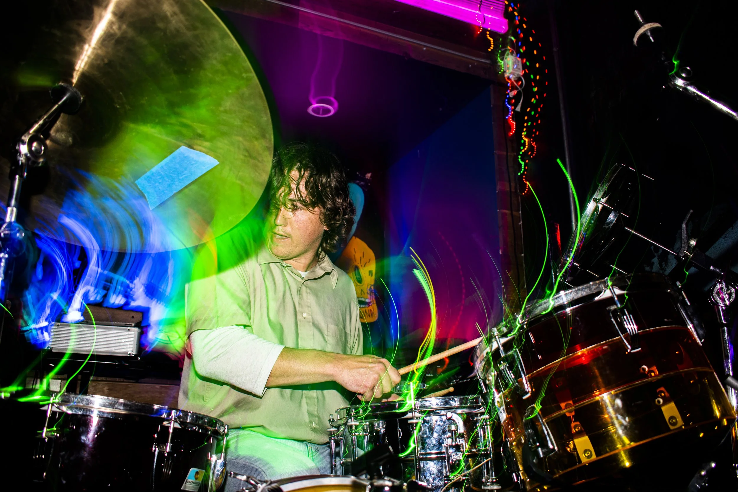 A man playing drums in a dimly lit room with colorful, moving light effects creating streaks of green, blue, and purple around him.