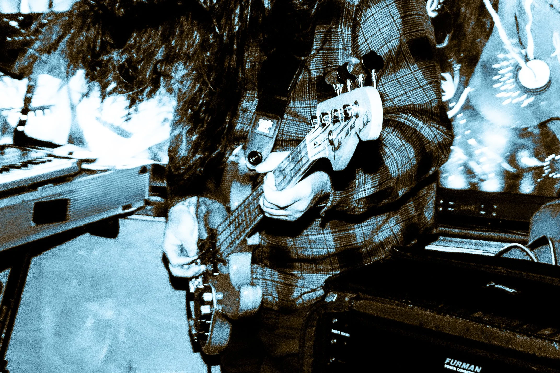 Close-up of a person wearing a plaid shirt playing an electric guitar in a music studio, with keyboard and amplifier visible.