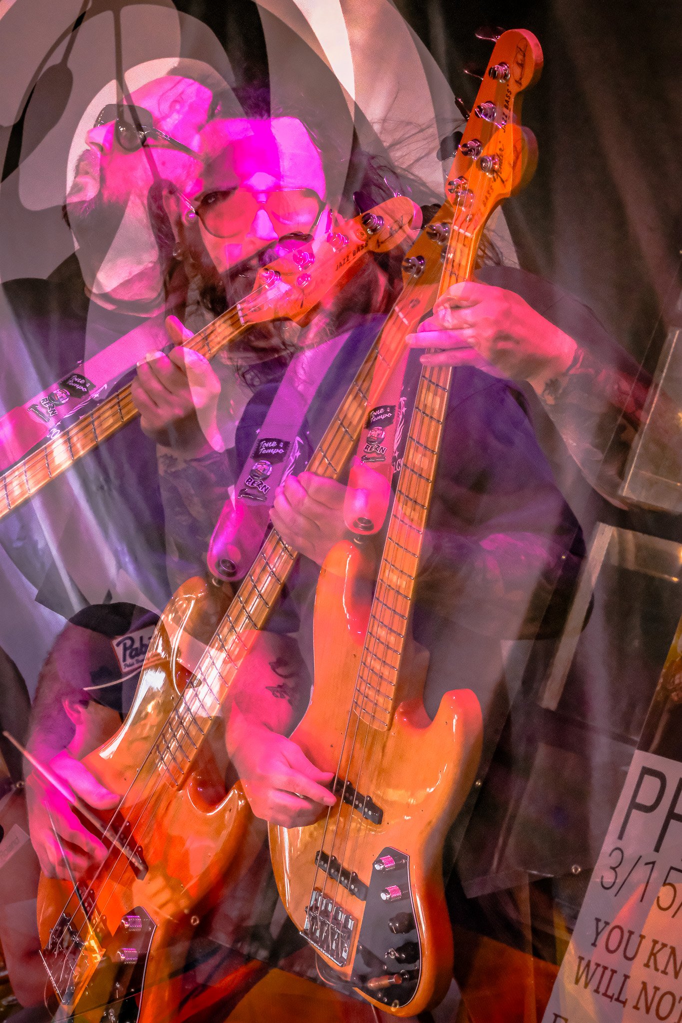 Tysonious Mink of the band Liquid Pennies playing a bass guitar, captured through a glass reflection with multiple layers and pink lighting.