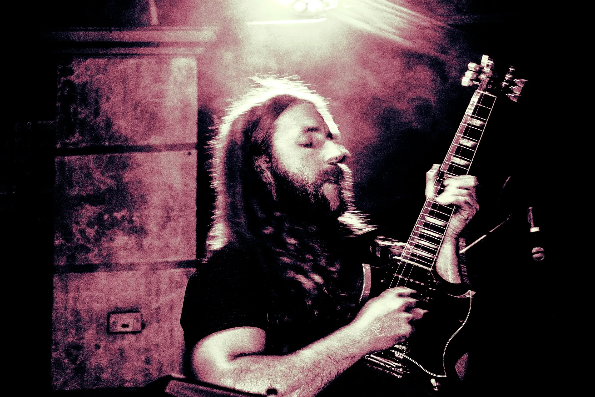 A man with long hair and a beard playing an electric guitar with eyes closed, illuminated by pinkish stage lighting.
