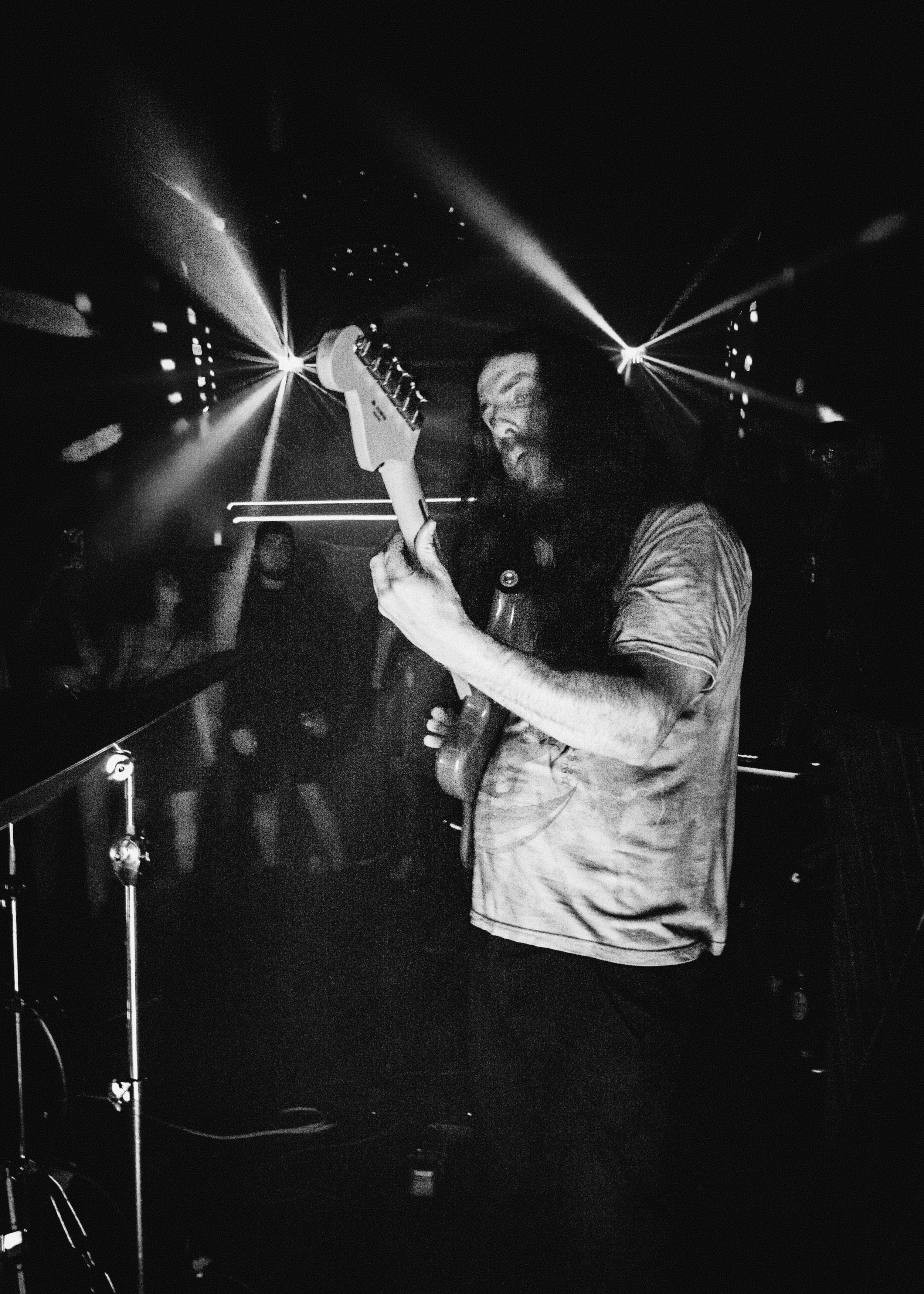 A man with long hair and a beard playing an electric guitar on stage under spotlights at a concert or nightclub.