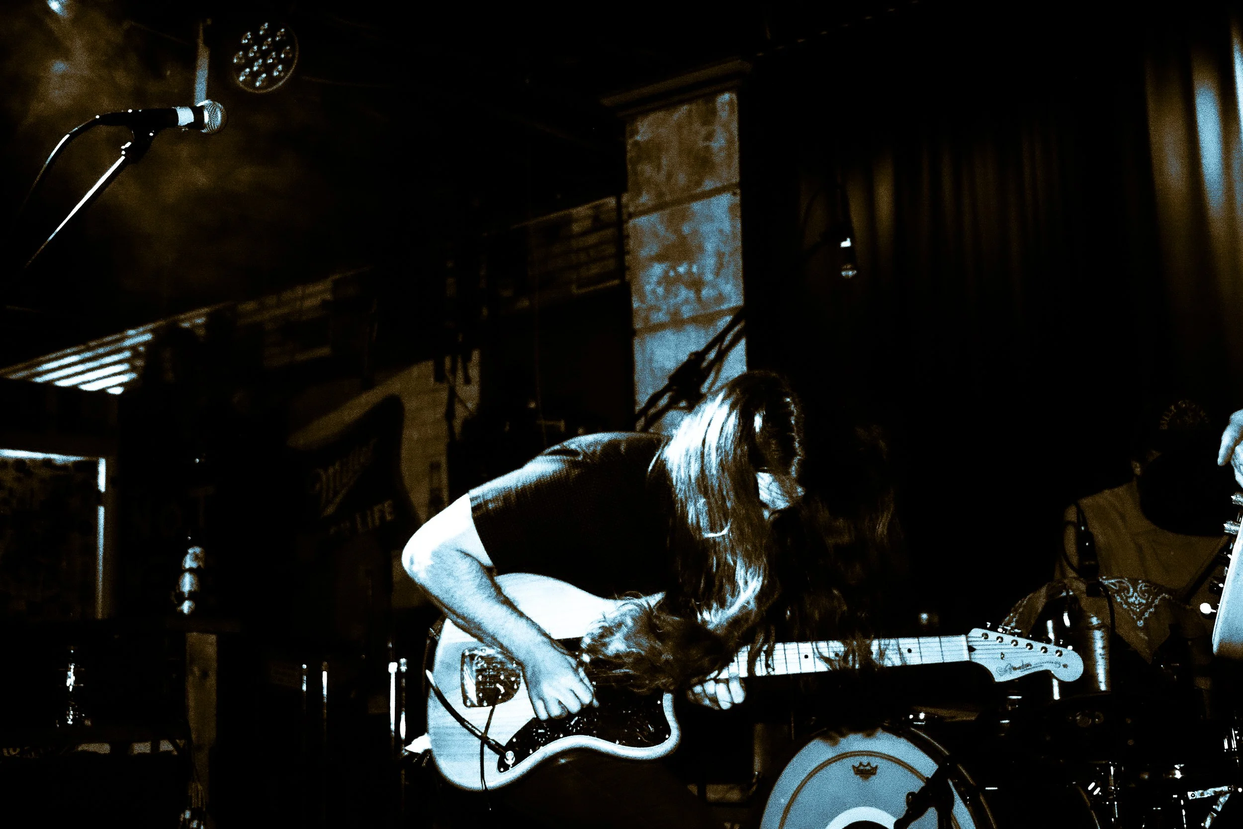 Musician with long hair playing an electric guitar on stage with a microphone stand nearby, dimly lit with warm, blue lighting.