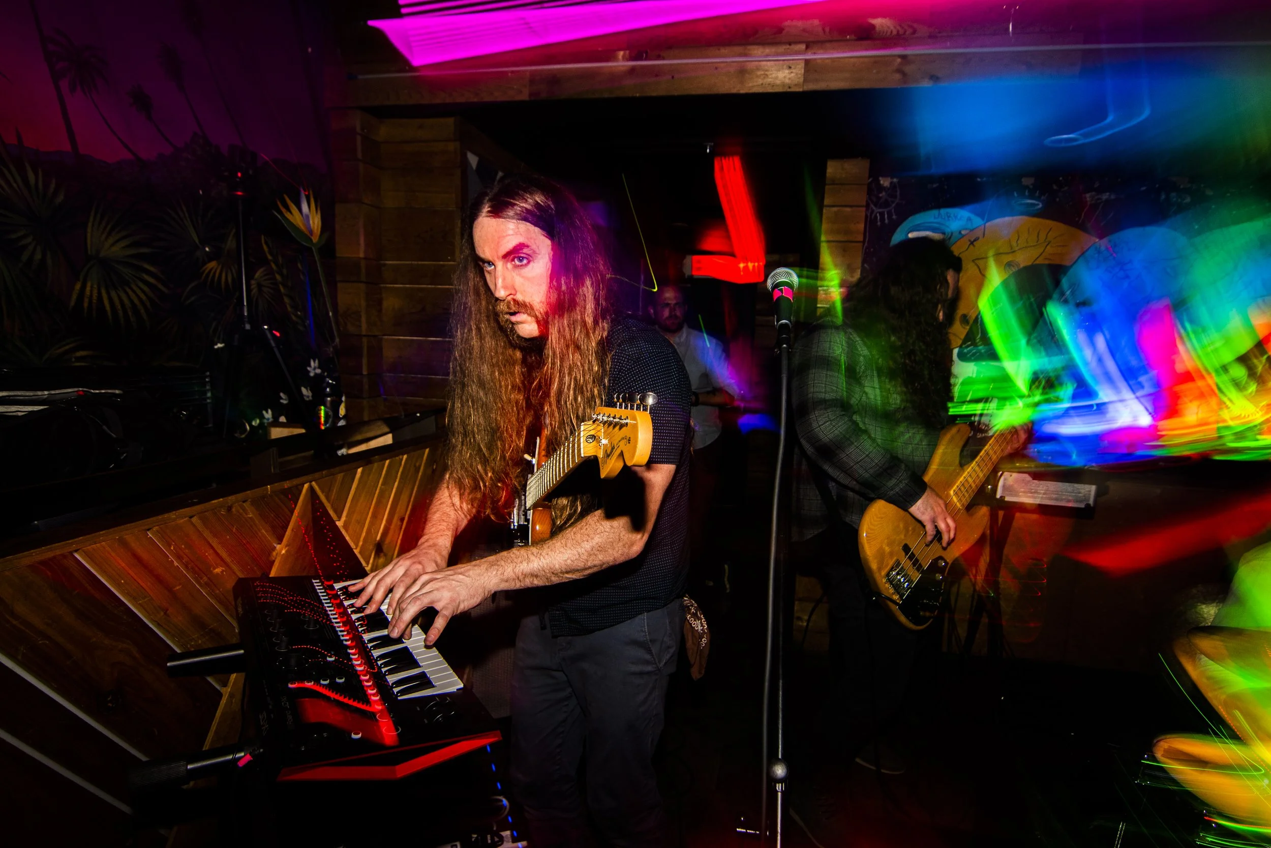 Musicians performing live at a venue with colorful light streaks. One musician is playing a keyboard with a guitar around his neck, and another is playing a bass guitar in the background. The scene is vibrant and energetic.