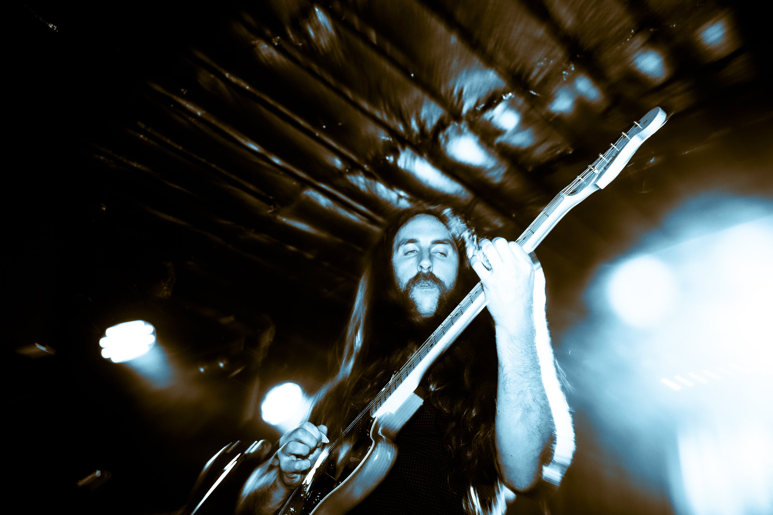 Rock band Liquid Pennies frontman/guitarist Chas BInns playing an electric guitar on stage under colorful lights.