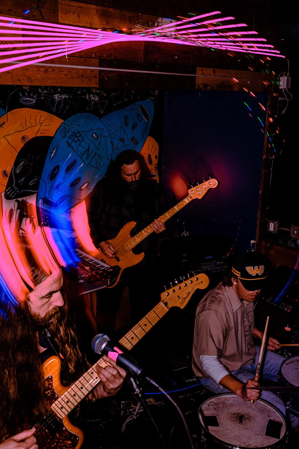 A band performing with two guitarists and a drummer in a colorful, neon-lit setting, featuring abstract artwork of faces on the wall behind them.