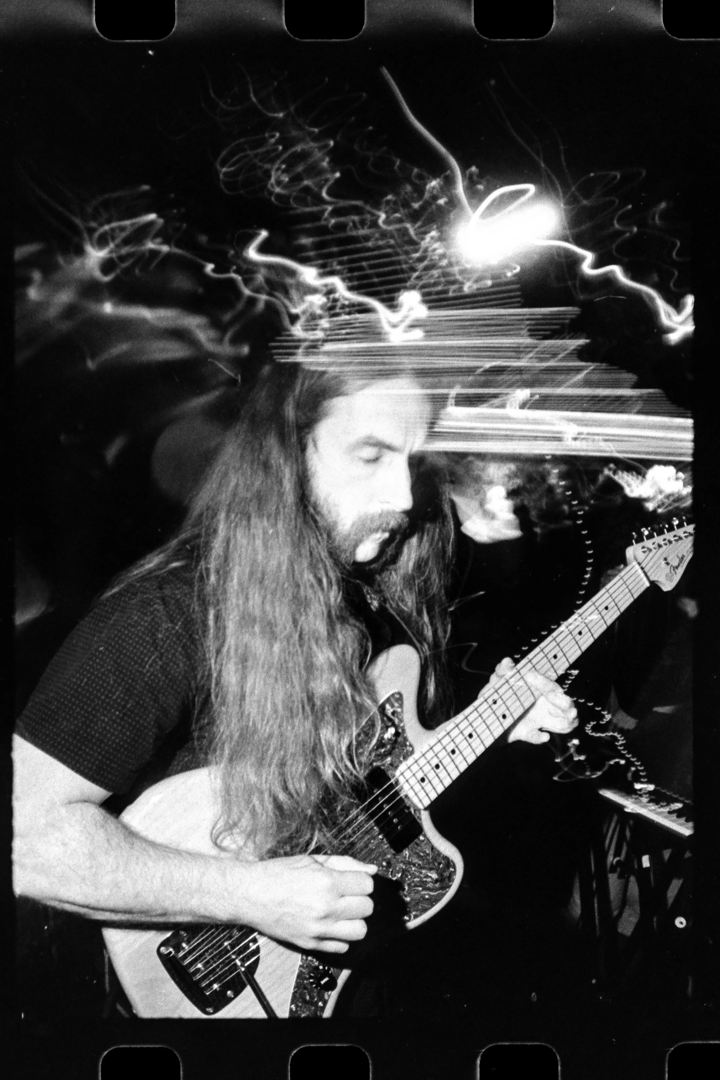 Black and white photo of Liquid Pennies frontman/guitarist Chas Binnsplaying an electric guitar with light streaks and motion blur in the background. Taken on a film camera.