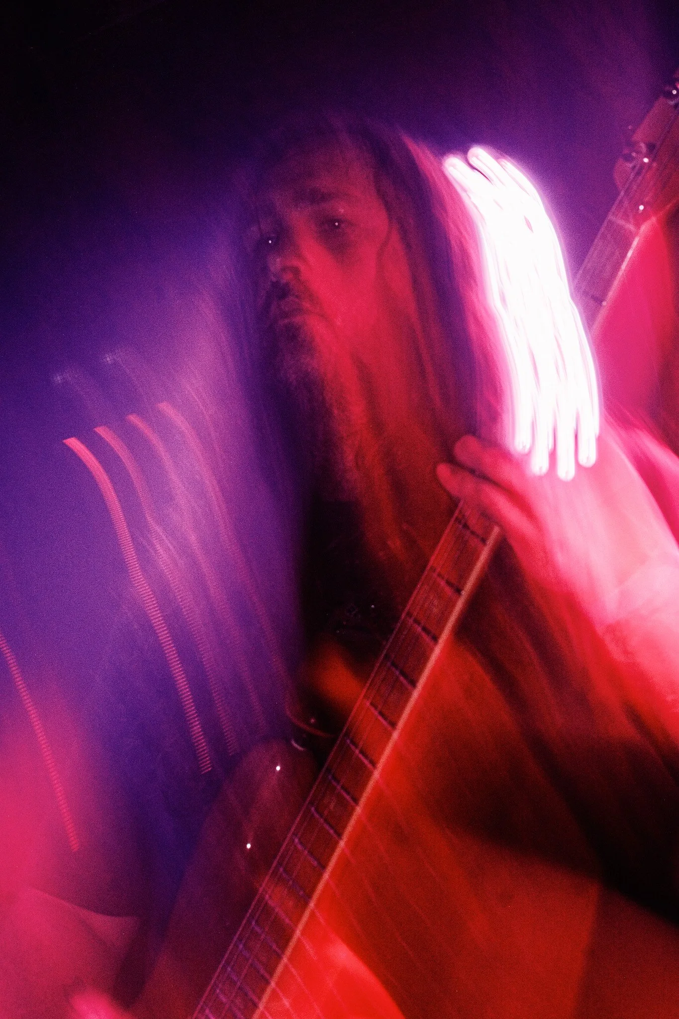 Tysonious Mink of the hard rock band Liquid Pennies playing a Fender Jazz Bass in a dimly lit setting with pink and purple neon lights, creating a foggy, artistic effect.