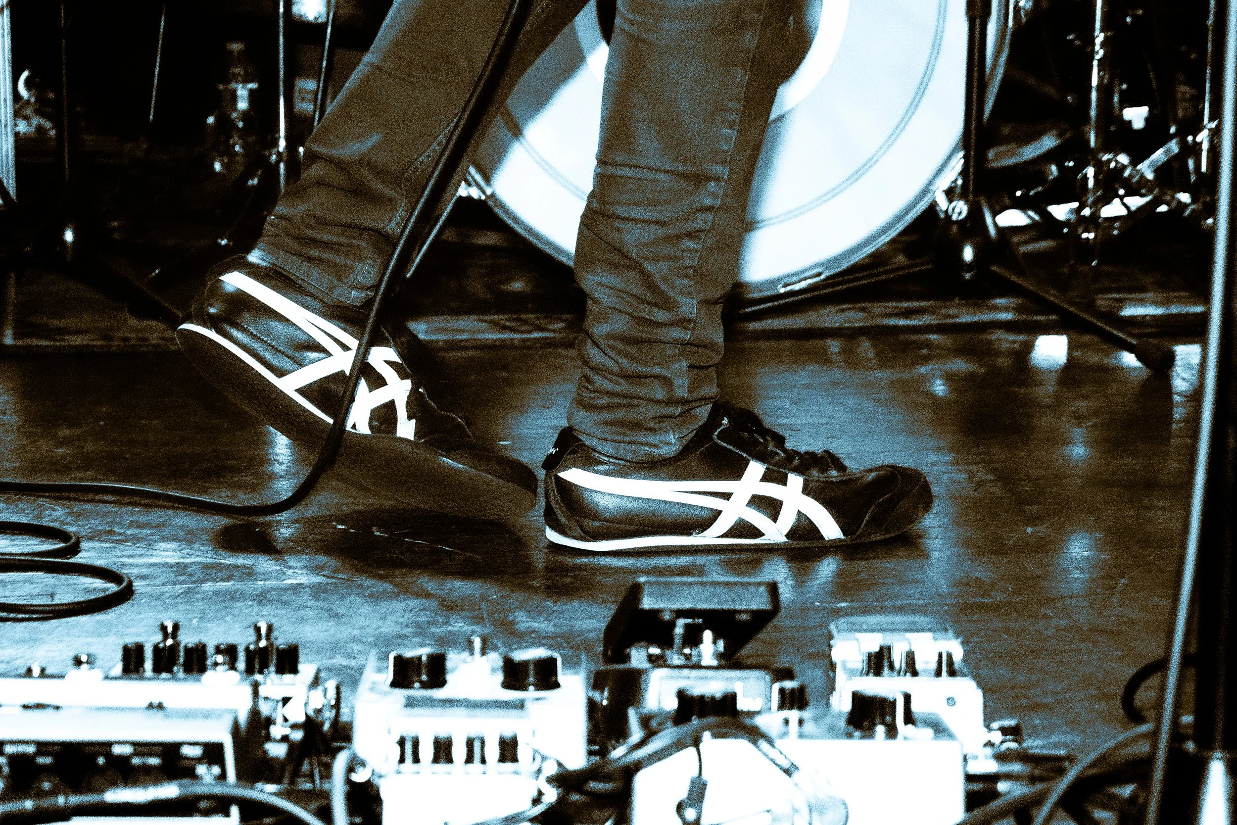Close-up of Rock band Liquid Pennies frontman/guitarist Chas BInns' feet wearing black athletic Onitsuka Tiger shoes with white reflective stripes, standing on a wooden floor among musical equipment and cables, with a drum kit in the background.