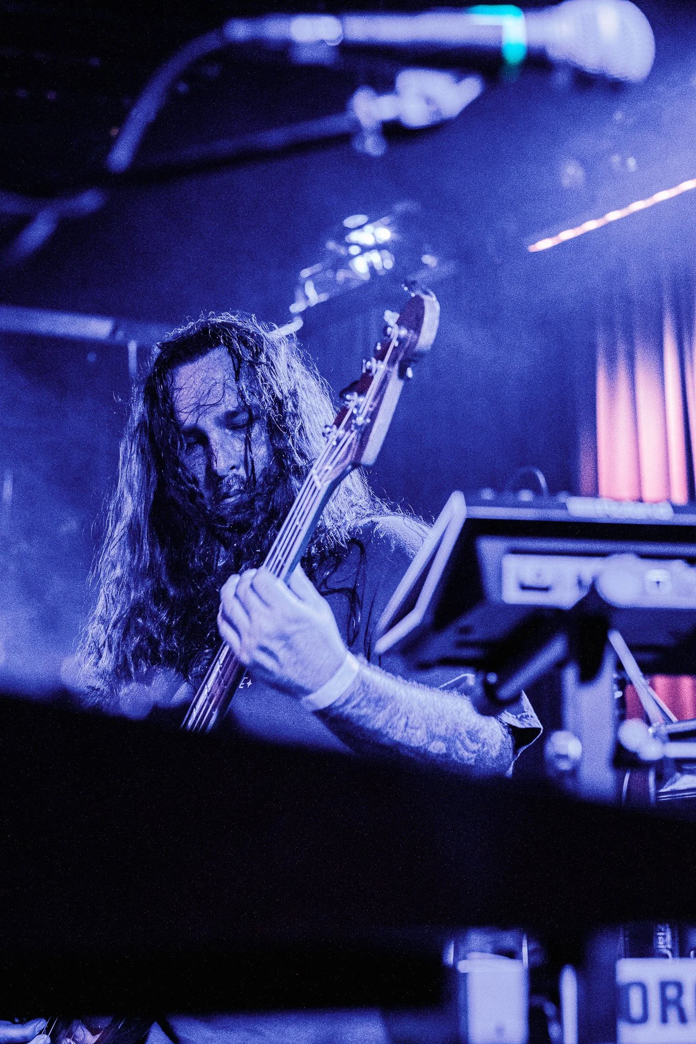 A musician with long curly hair, playing an electric bass guitar on stage with blue lighting.